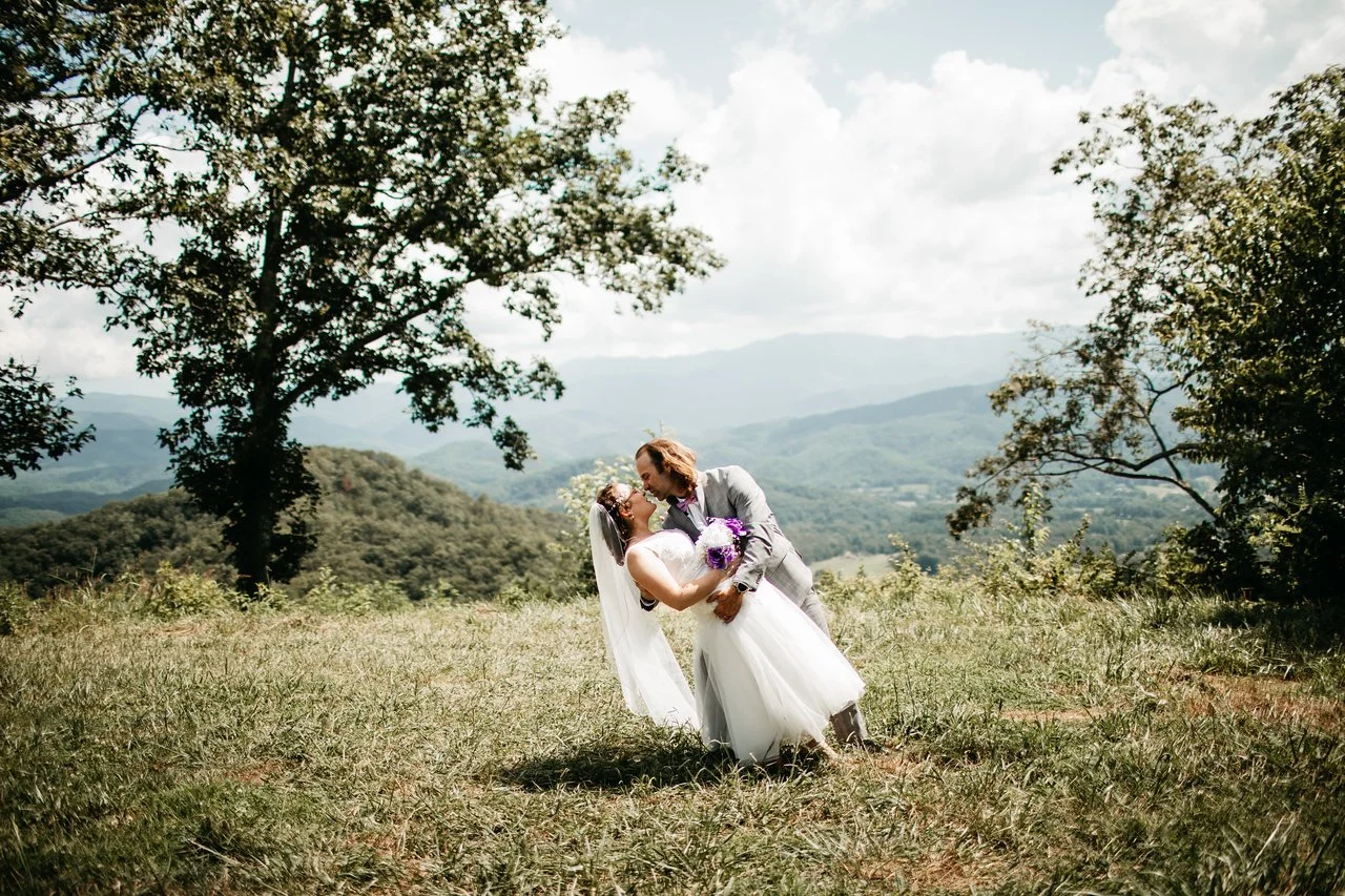 The couple perform a leaning kiss in a clearing in the woods, overlooking green mountains in the distance behind them.