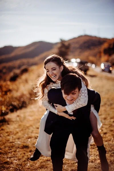 The groom playfully gives the bride a piggyback ride. They are both laughing. In the distance there are a few cars, and beside the road, a hill slopes down into a forest below.