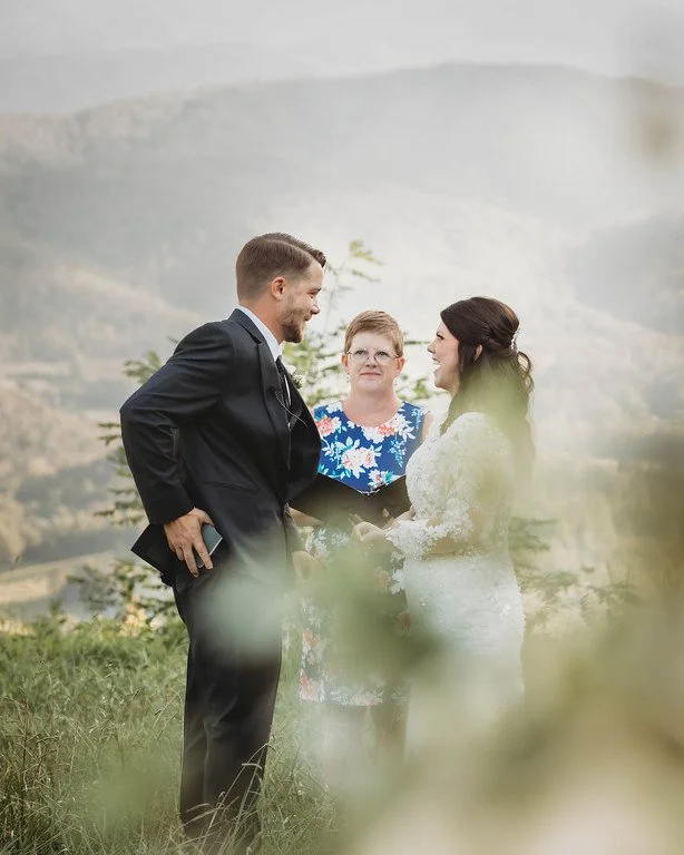 The couple stand facing eachother in front of the officiant, while one puts their vows back in their pocket. They are slightly obscured by wildflowers in the shot, and behind them is a lush, green mountain.
