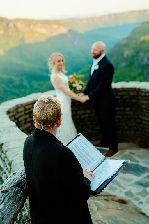 The couple stand at a small overlook, facing eachother in the background. In the foreground stands the officiant watching the couple at the altar. Beyond the overlook is a deep green valley.