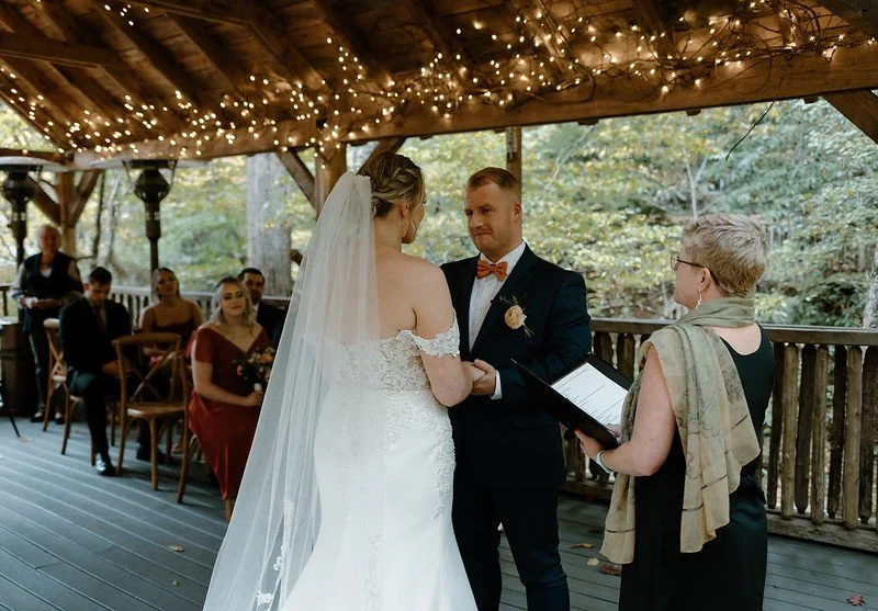 The couple face eachother at the altar, at a 3/4 angle to the camera. The officiant is seen on the right side of the shot. In the background are wedding guests and the wall of the pavilion can be seen beyond that, with sparkling string lights.