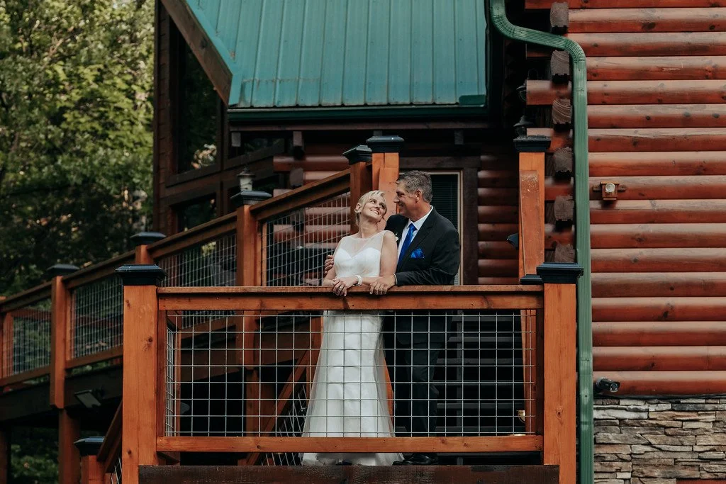 The couple stand side by side, sharing a glance, on the porch of a cabin. The green metal roof and large windows of the cabin can be seen behind them, and beyond that, peeks a bright green forest.