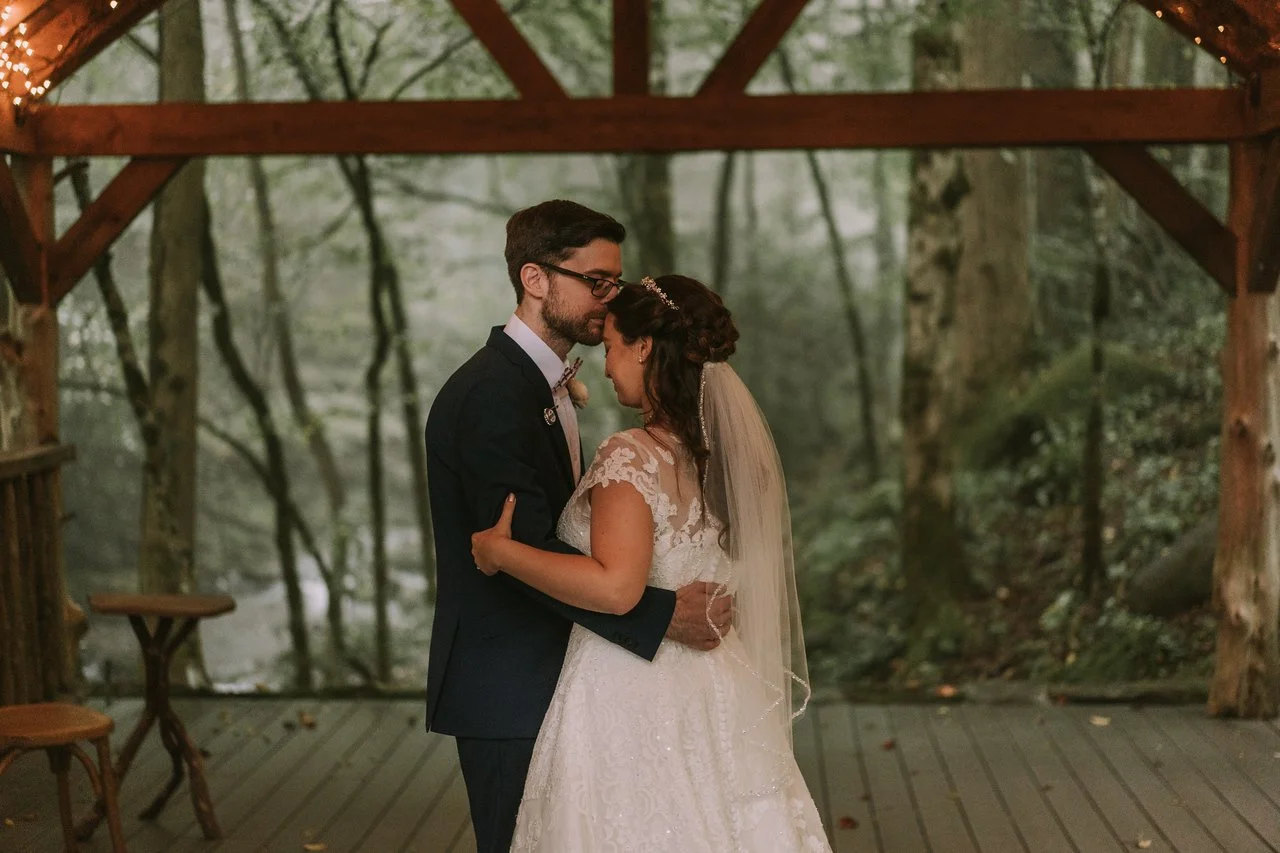 The couple stand alone. slow dancing the wedding pavilion. The edge of the pavilion overlooks flowing river, and a dense green forest.