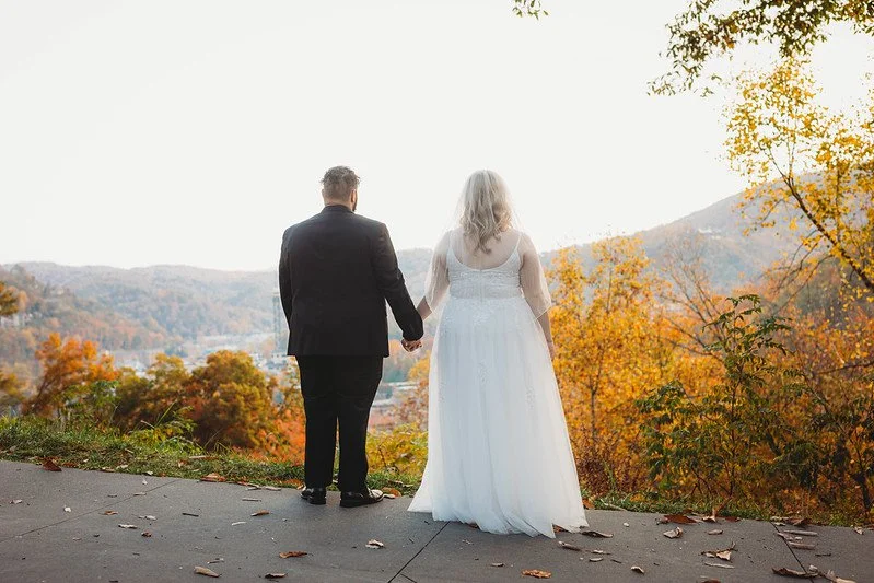 The couple stand with their backs to the camera, overlooking Gatlinburg during autumn. The trees nearest to them are yellow, and the mountain in the distance is dotted with many colors of leaves.