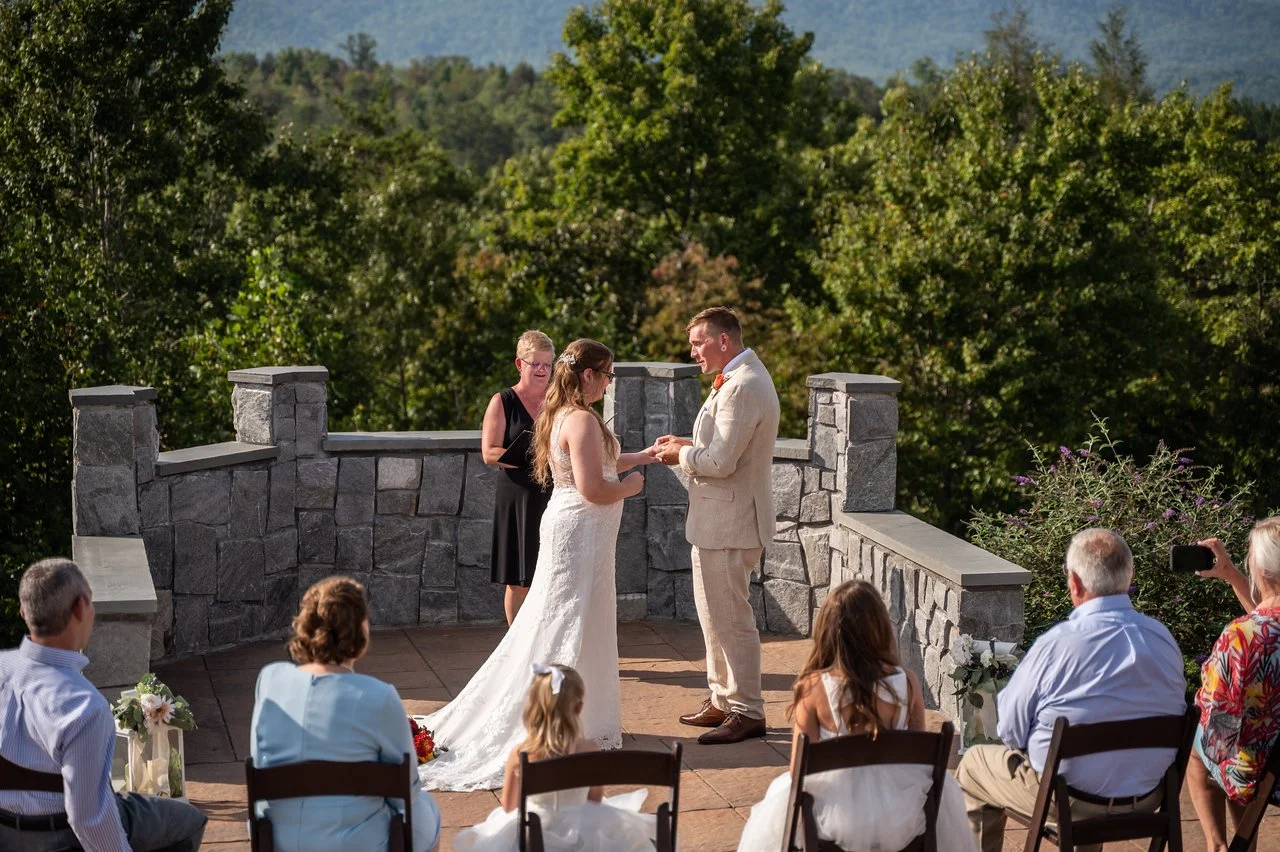 The couple stand facing eachother at the altar while one puts a ring on the other. Beside them is the officiant. In the foreground, wedding guests sit in wooden chairs. Behind the officiant is a stone wall overlooking a green forest.