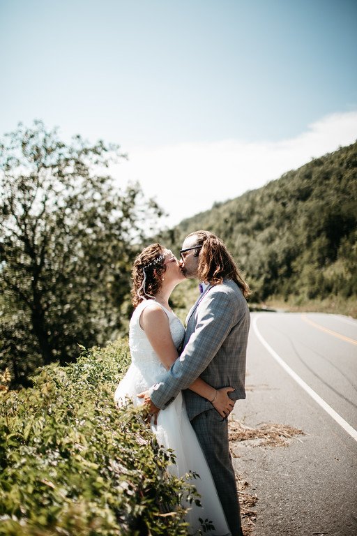 The couple stand, embracing eachother by the butt, at the side of the road, kissing. Behind them is a mountain and a slightly cloudy blue sky.