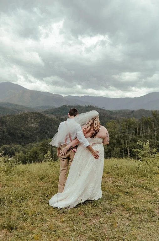 The couple stand side by side, facing away from the camera. They each have a hand on the other's butt as they overlook the lush green forest and tall mountains in the distance. The sky is overcast.