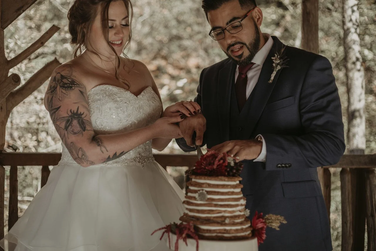 The couple stand in the wedding pavilion, ceremoniously cutting their unique layered wedding cake, smiling.