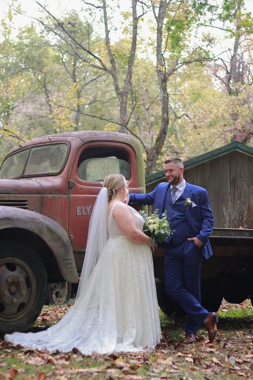 The couple lean against an antique red truck, facing eachother. In the background there is an old wooden building and a forest behind it. The truck has the words "Ely's Mill" painted on it, partially obscured by the couple.
