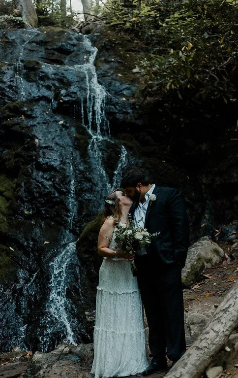 The couple stands in the woods, embracing and sharing a kiss. A waterfall trickles down behind them.
