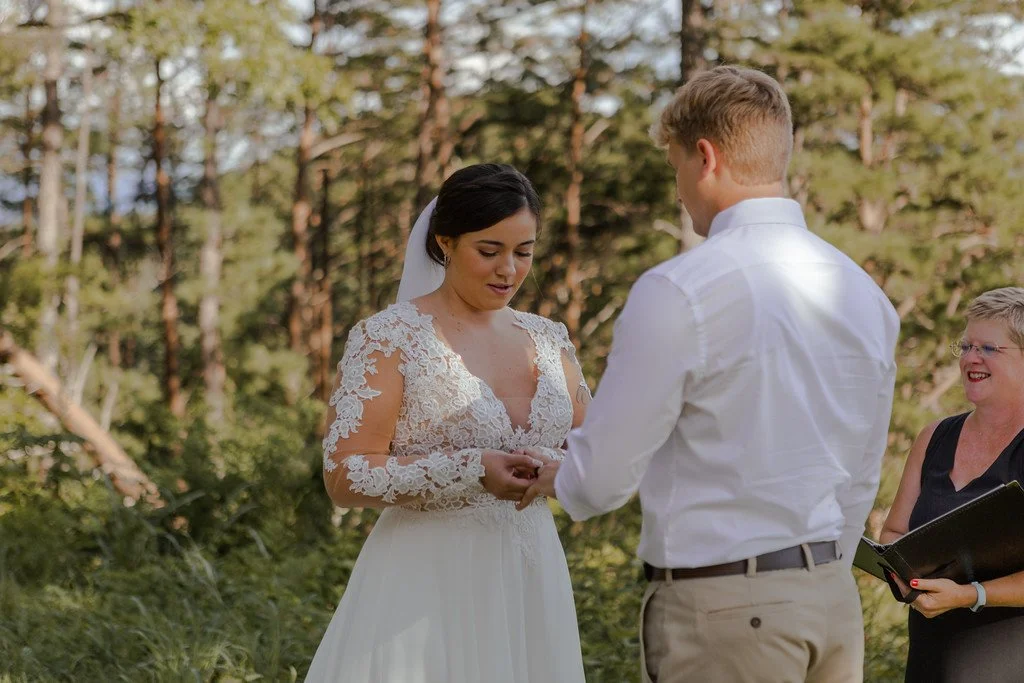 The couple stands at a 3/4 view to the camera, while one puts a ring on the other. In the background are many tall evergreen trees, and the sun is moderately low in the sky out of frame.