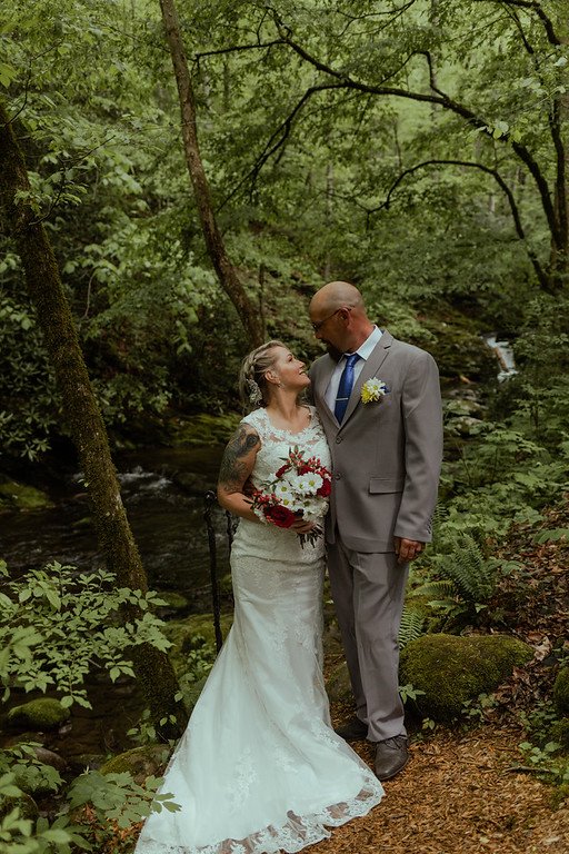 The couple stands side by side, looking at eachother, embracing. In the background, there is a flowing stream, lush vegetation, and a bright green forest.