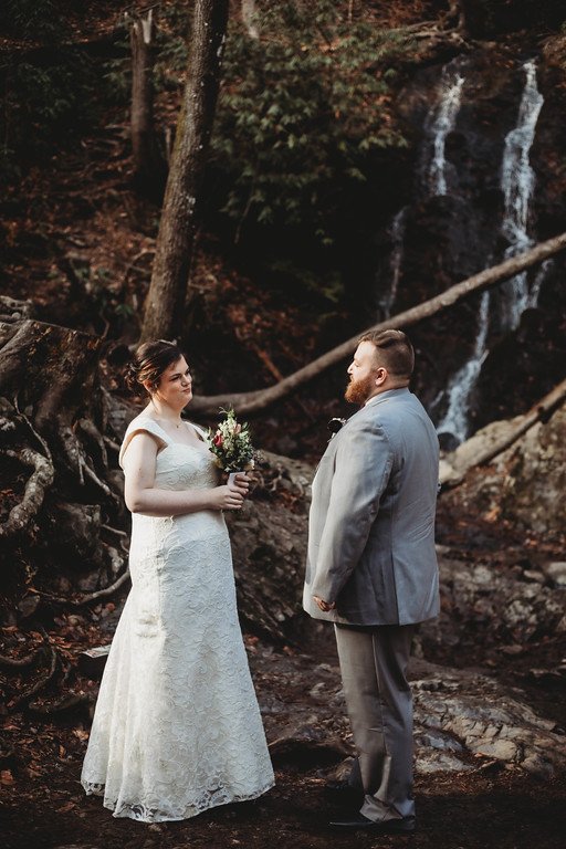 The couple stands in the woods, facing eachother. Behind them is a trickling waterfall and some trees.