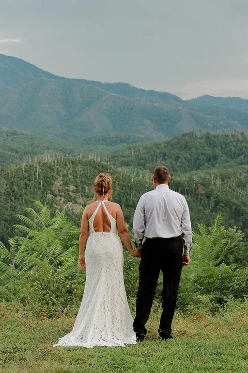 The couple stand facing away from the camera, holding hands. They overlook the smoky mountains in the distance, and green vegetation in front of them.