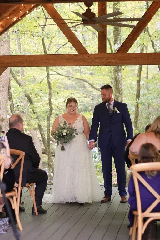 The couple holds hands in a wooden pavilion facing the wedding guests seen in the foreground on either side of the aisle. In the background there is a bright green forest.