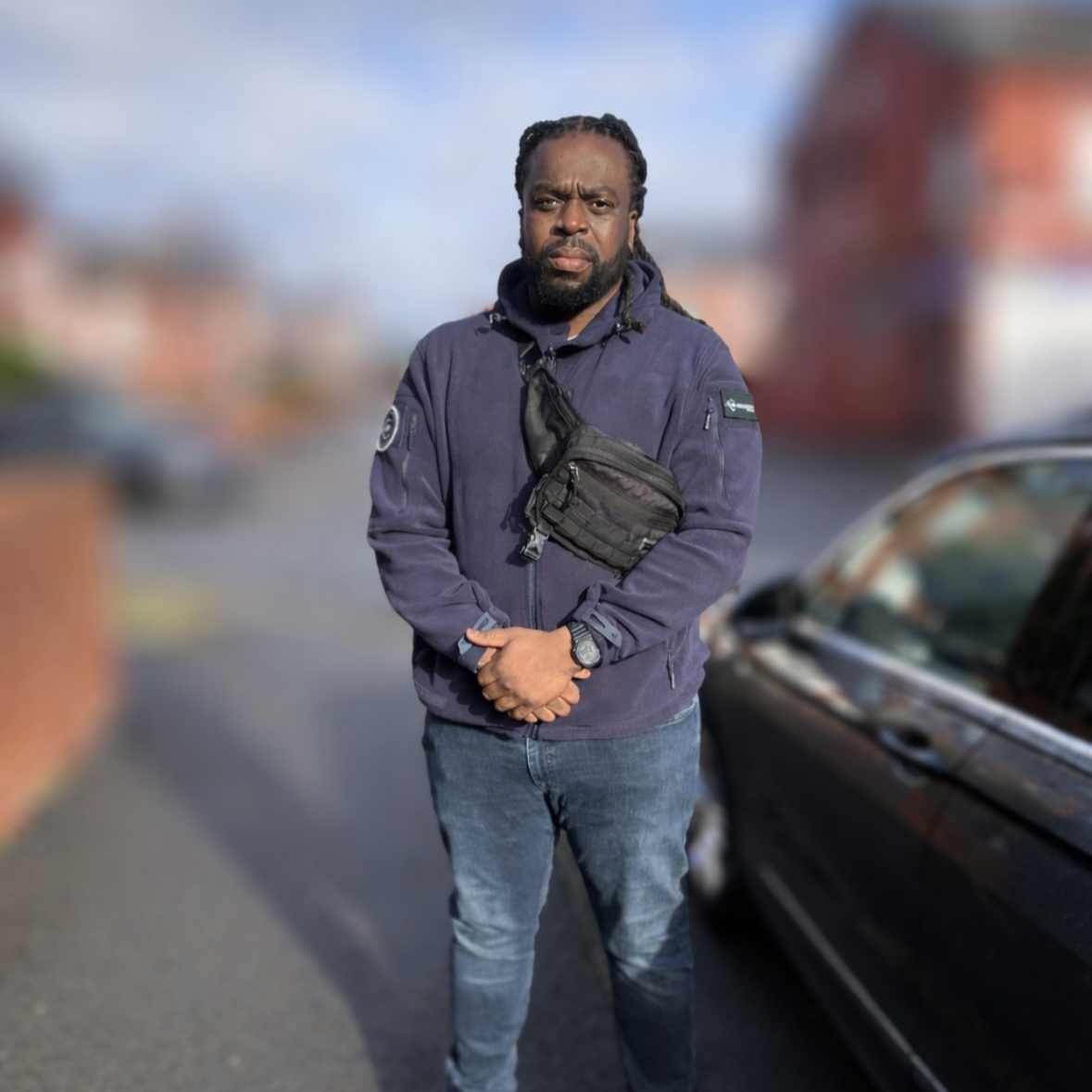 A man standing outdoors near a car, wearing a blue jacket and a crossbody bag, with a serious expression and long dreadlocks.