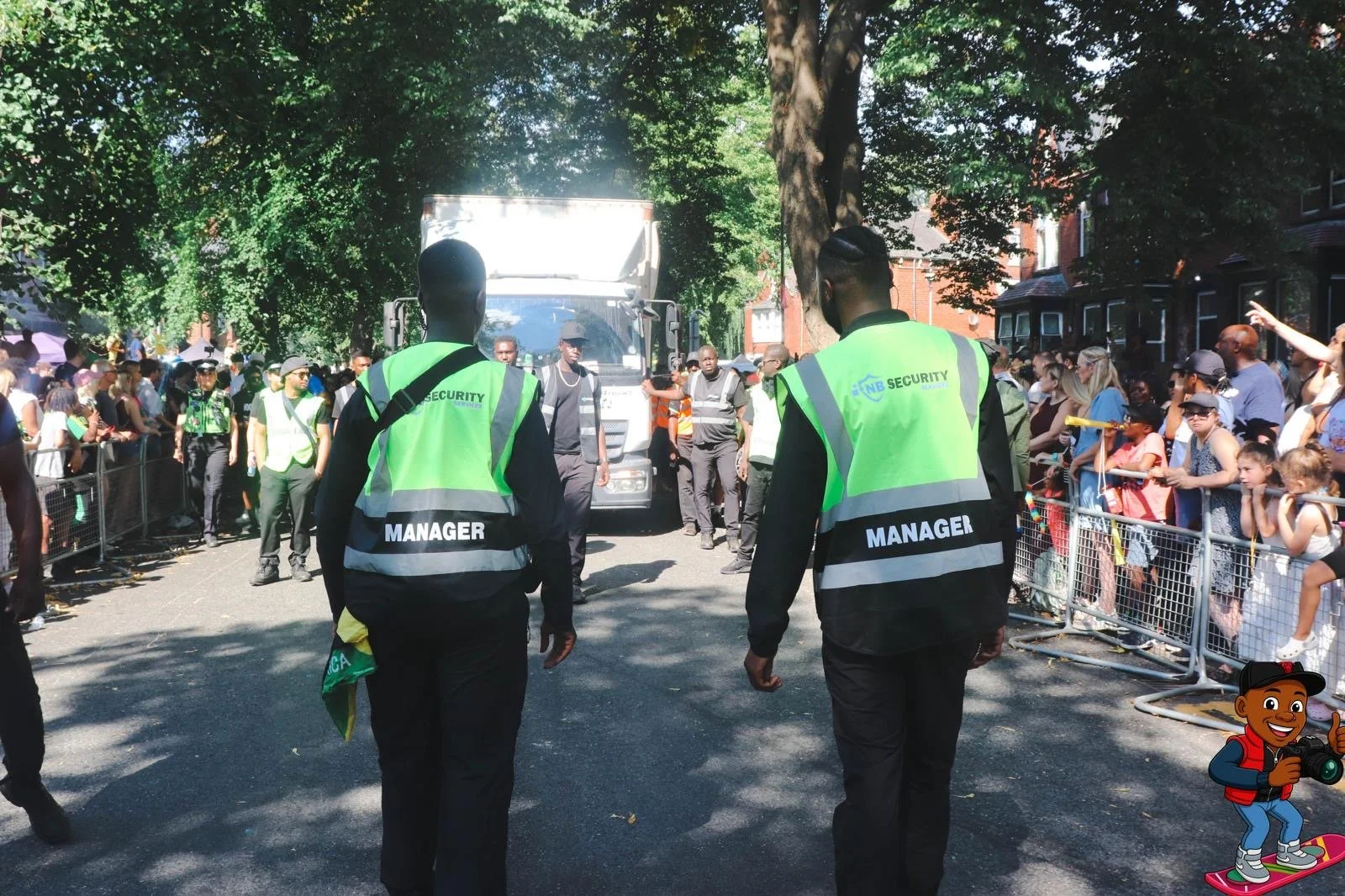 Crowd of people behind metal barriers on both sides of a street, attended by security personnel wearing neon green vests marked 'Security' and 'Manager'. In the background, a large white truck and tall trees with green leaves.