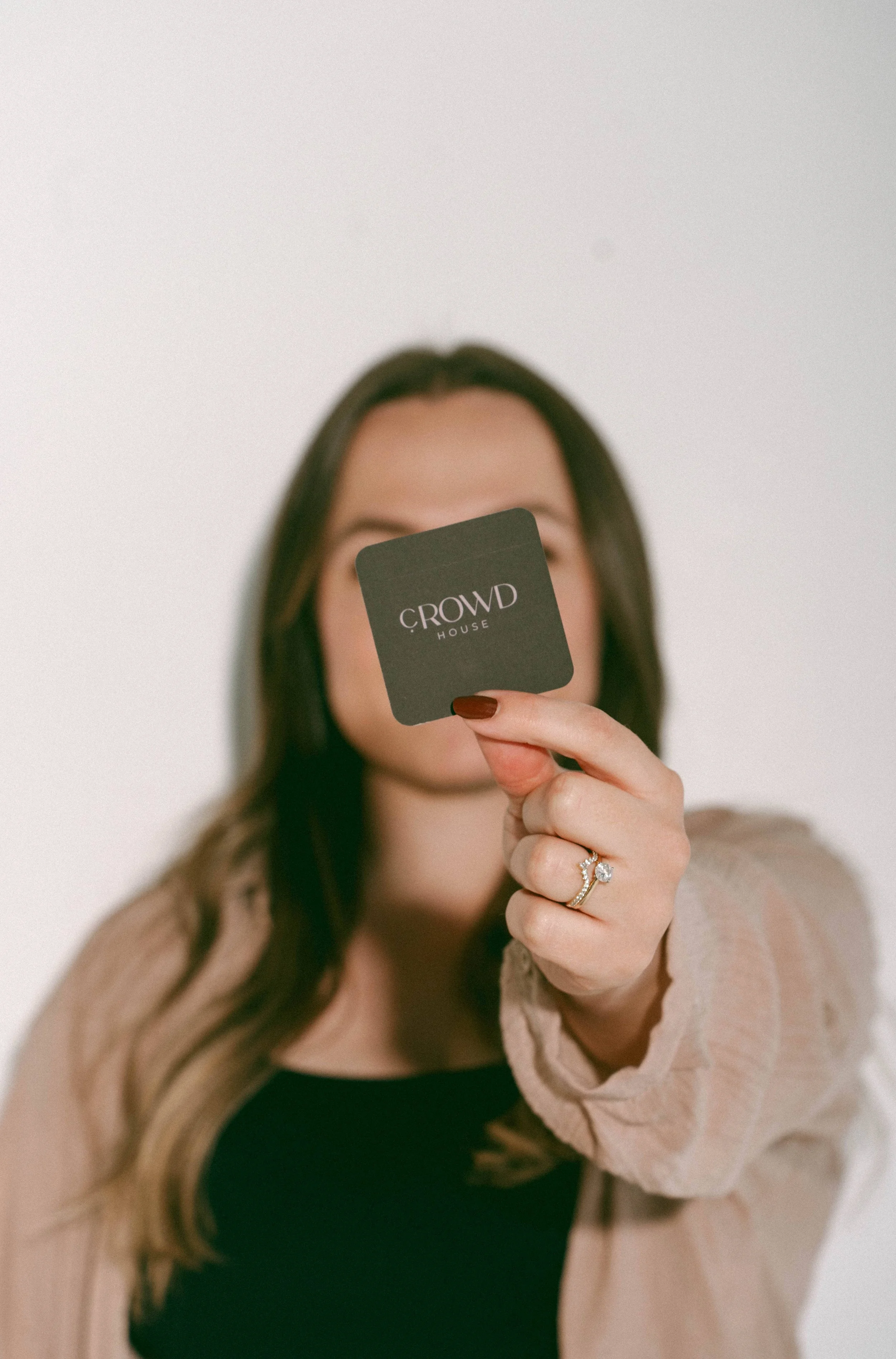 A woman holding up a square business card with C-rowd House's logo in front of her face.