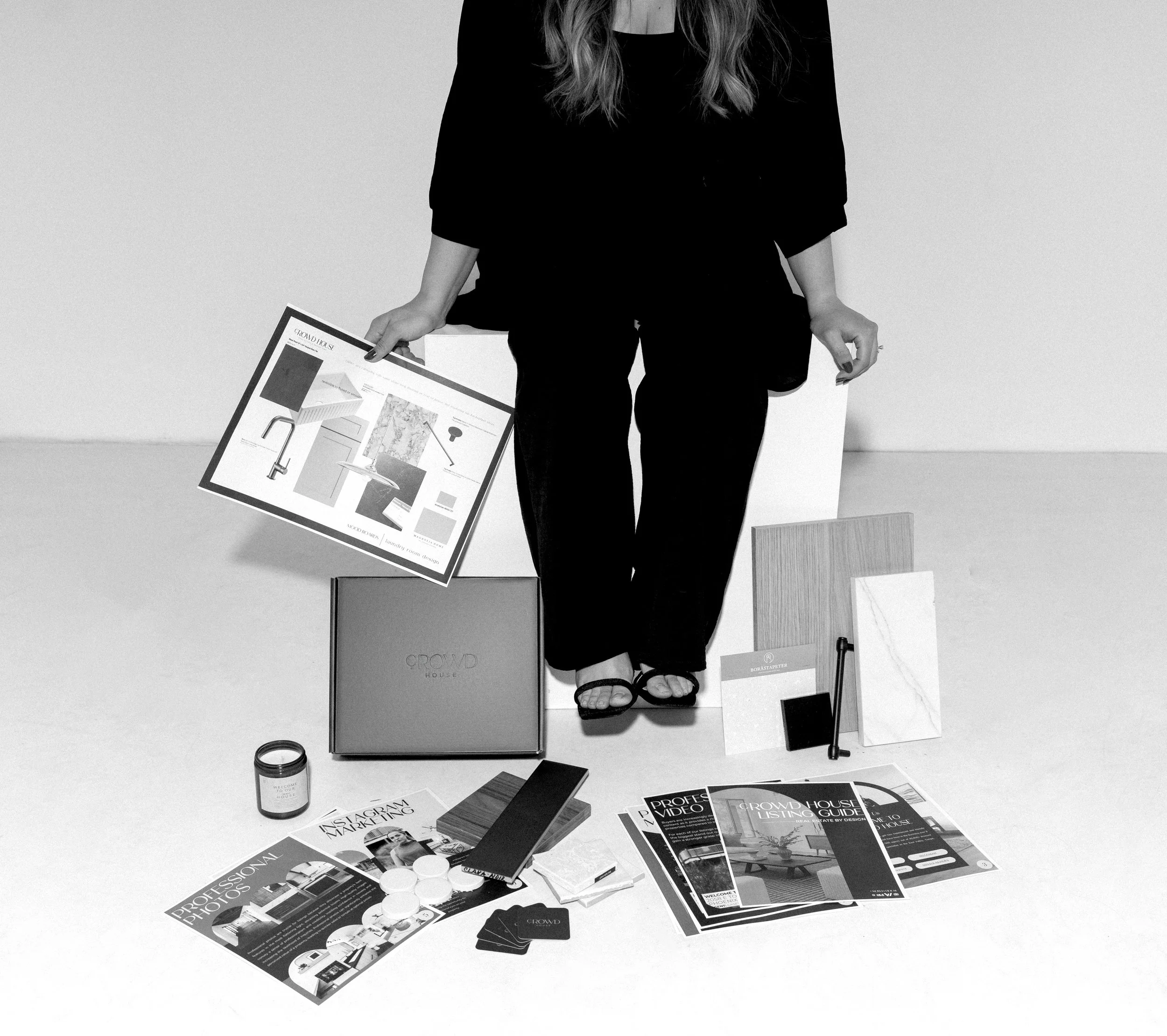 A woman sitting on a cube with various papers, packaging, and interior design samples on the floor around her.