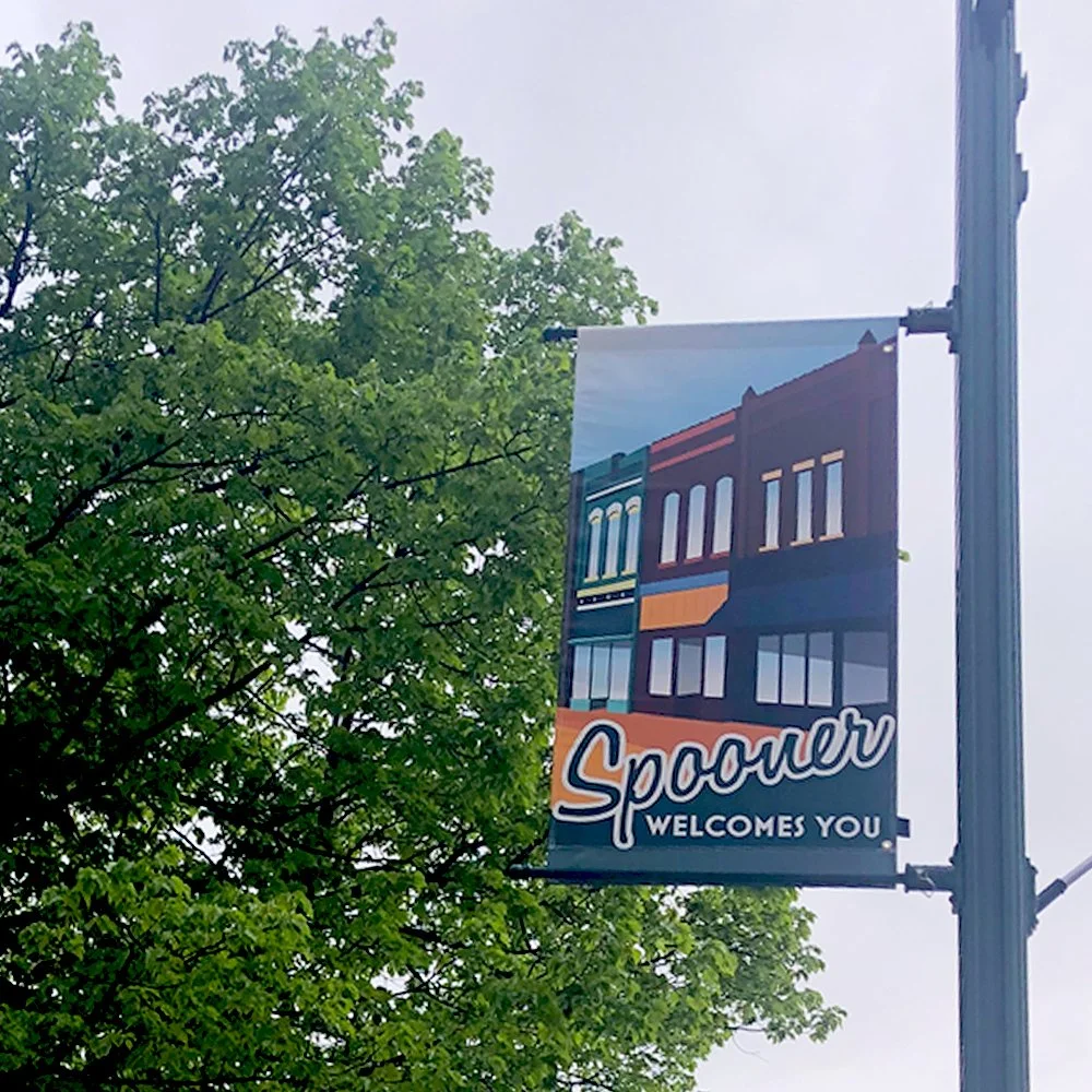Banner on lamp post reads "Spooner welcomes you" with illustration of historic downtonw