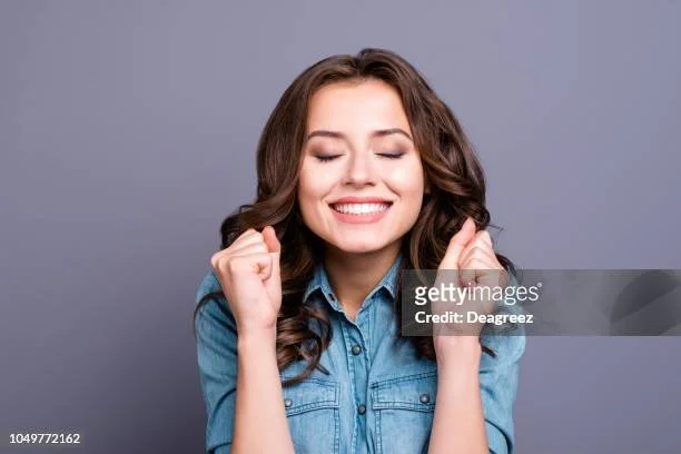 A young woman with brown hair wearing a denim shirt smiling and celebrating with her eyes closed.