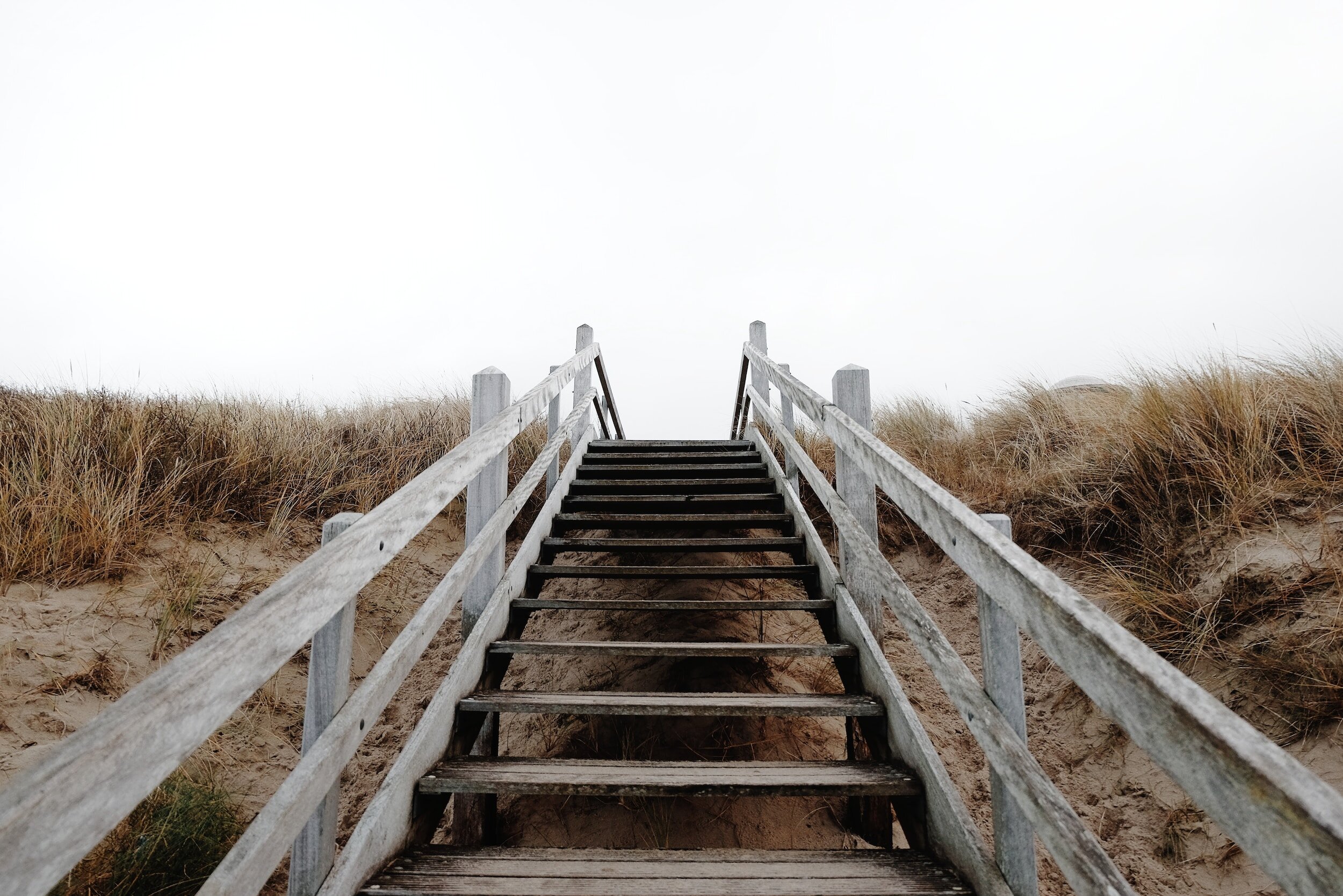 Wooden staircase leading up sandy dunes with tall grass on either side under a cloudy sky