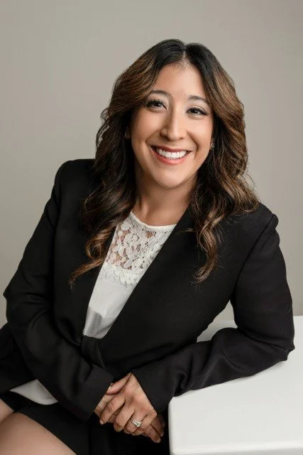 Portrait of a woman with wavy brown hair wearing a black blazer and a white lace top, smiling, against a plain neutral background.