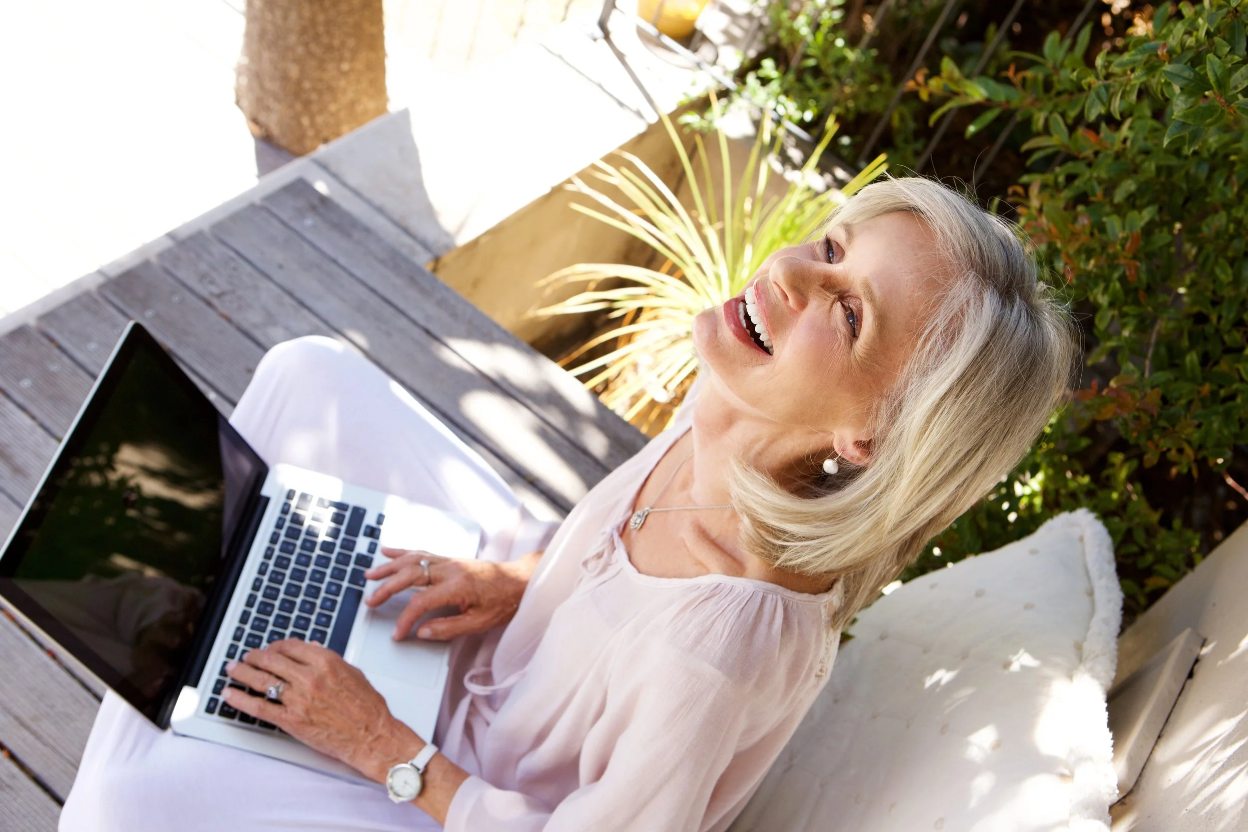 Older woman sitting outdoors on a patio, working on a laptop, smiling, surrounded by plants and sunlight.