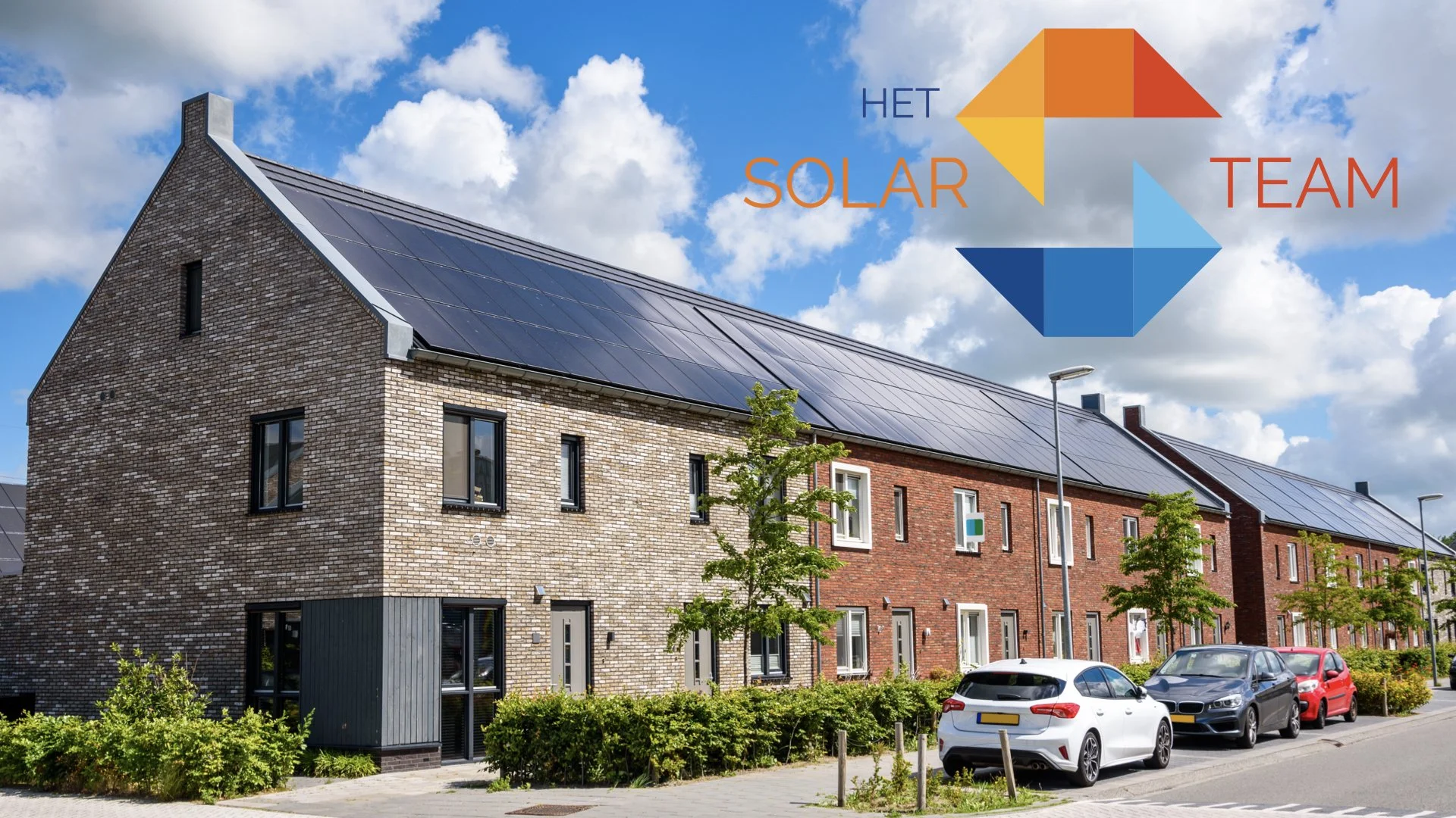 Residential neighborhood with modern brick houses topped with solar panels, parked cars, trees, and streetlights under a partly cloudy sky.