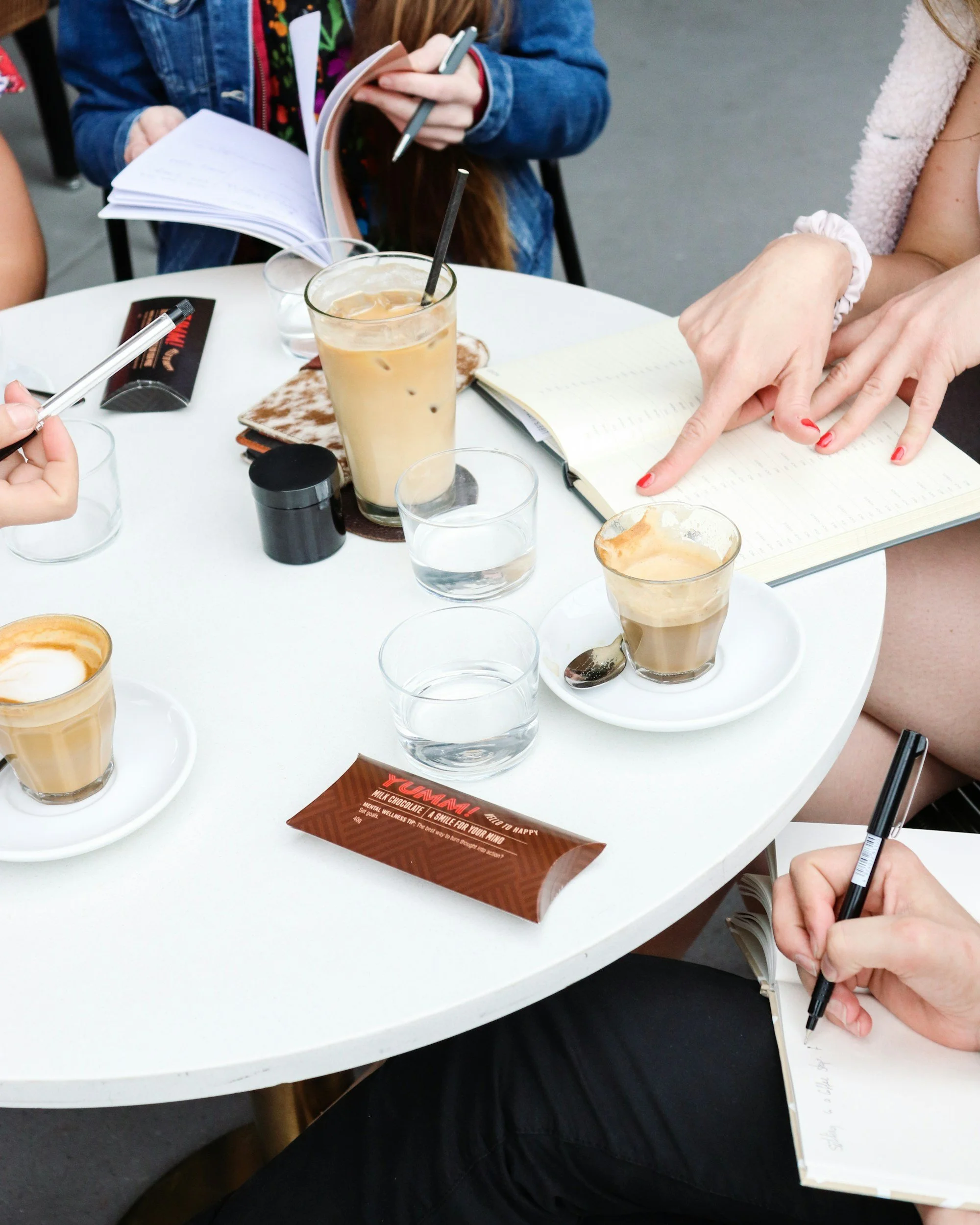 A group of people sitting around a white table having coffee, with notebooks and pens, engaged in a discussion.