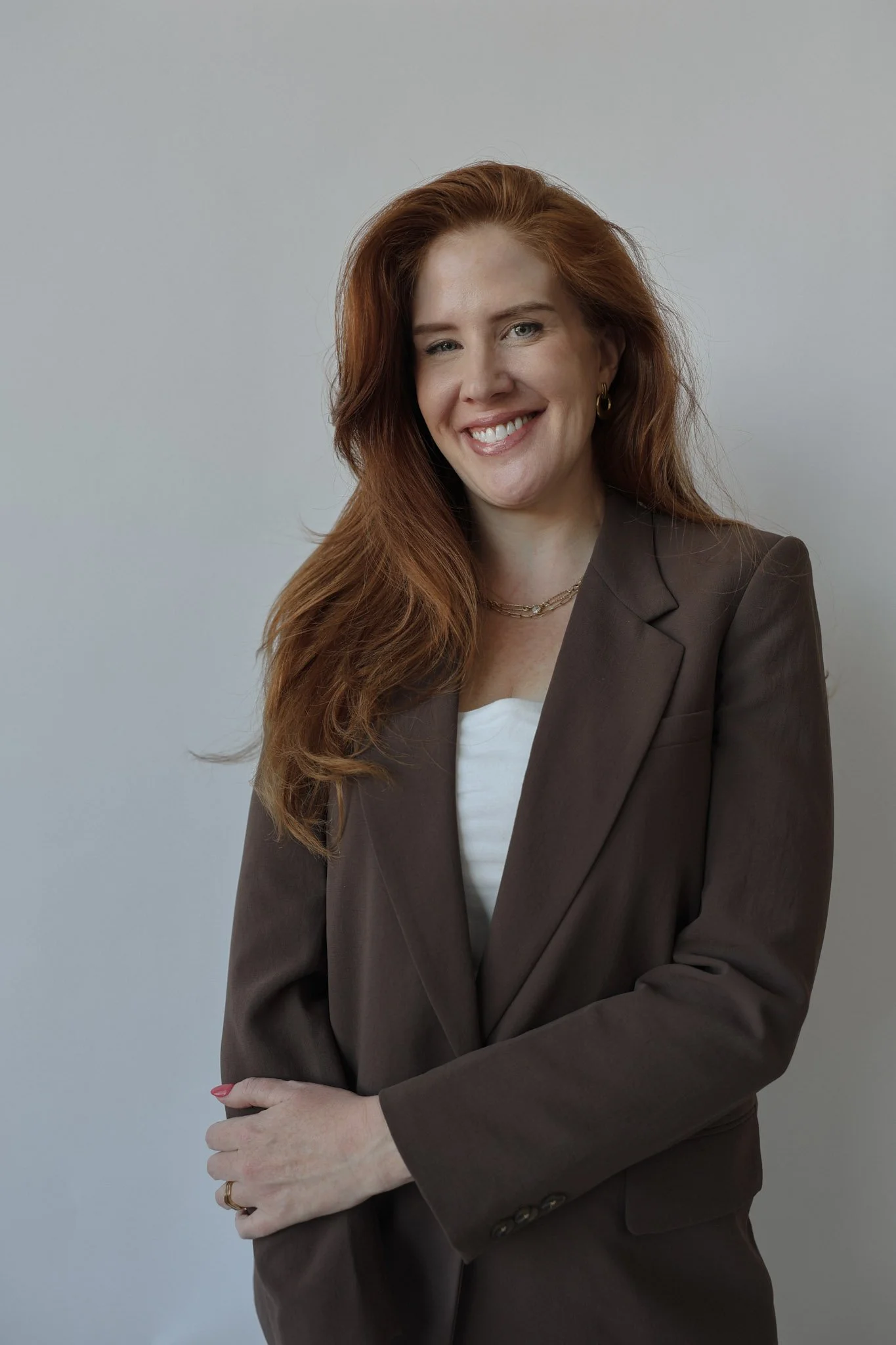 A woman with long red hair, wearing a brown blazer over a white top, smiling at the camera against a plain light gray background.