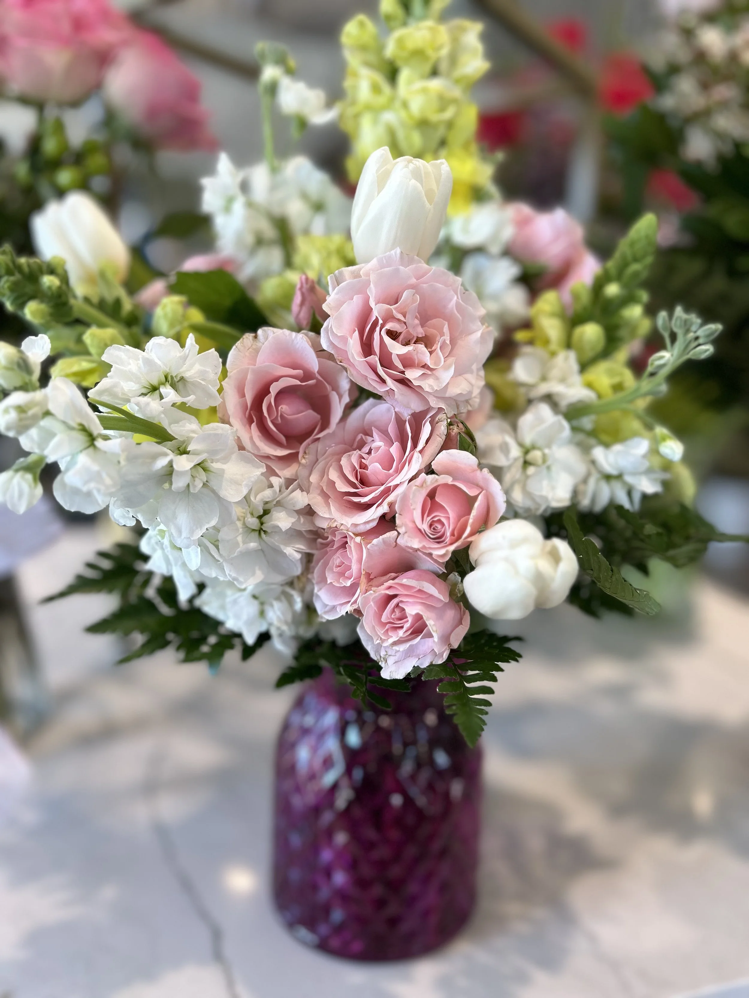 A floral arrangement in a gray vase on a woven basket, featuring white hydrangeas, pink roses, pink tulips, dark red flowers, white stock flowers, pink waxflowers, and greenery, set against a dark blue wall.