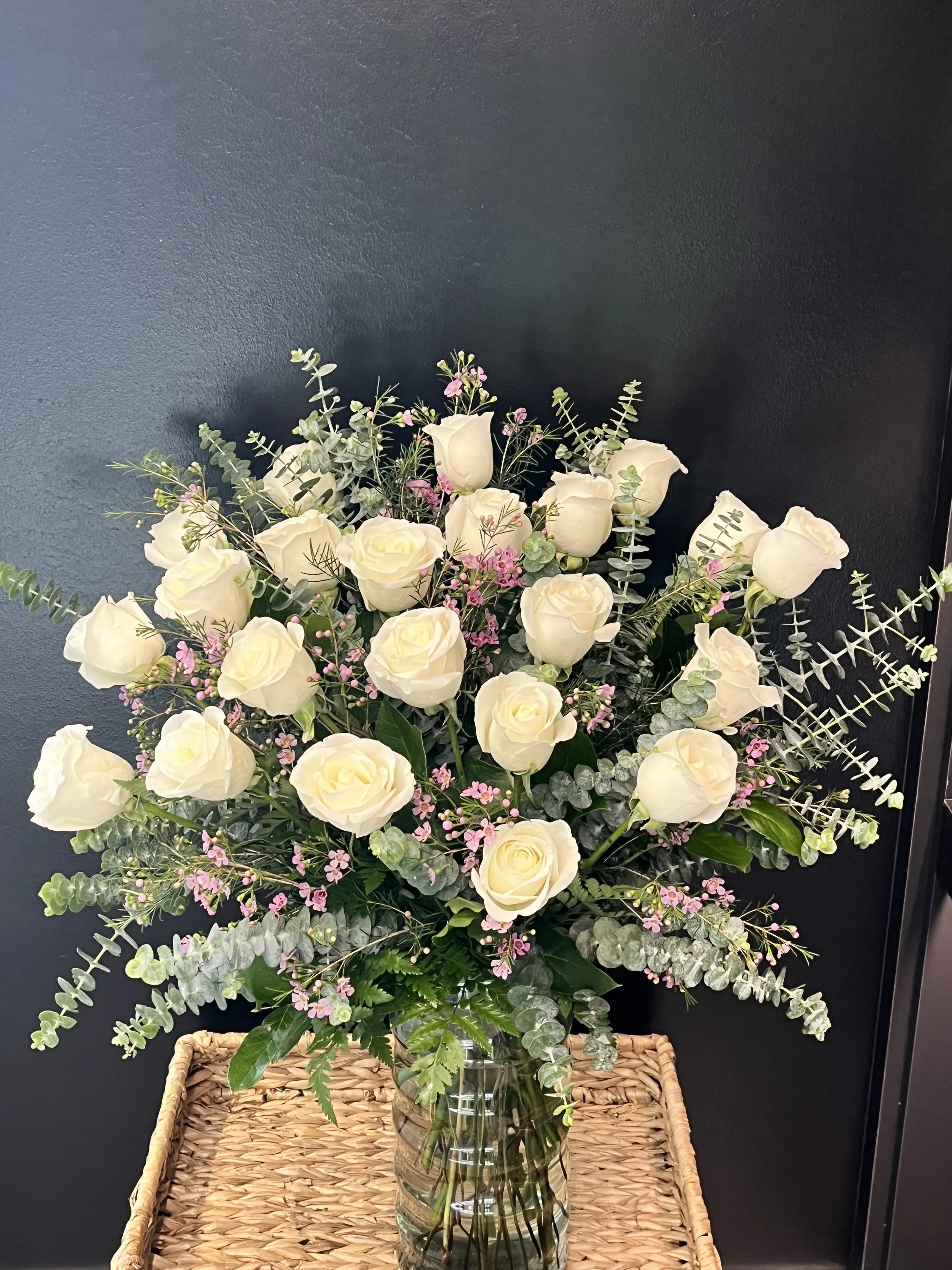 A large bouquet of white roses with pink and green filler flowers and greenery in a glass vase on a woven basket table against a dark background.