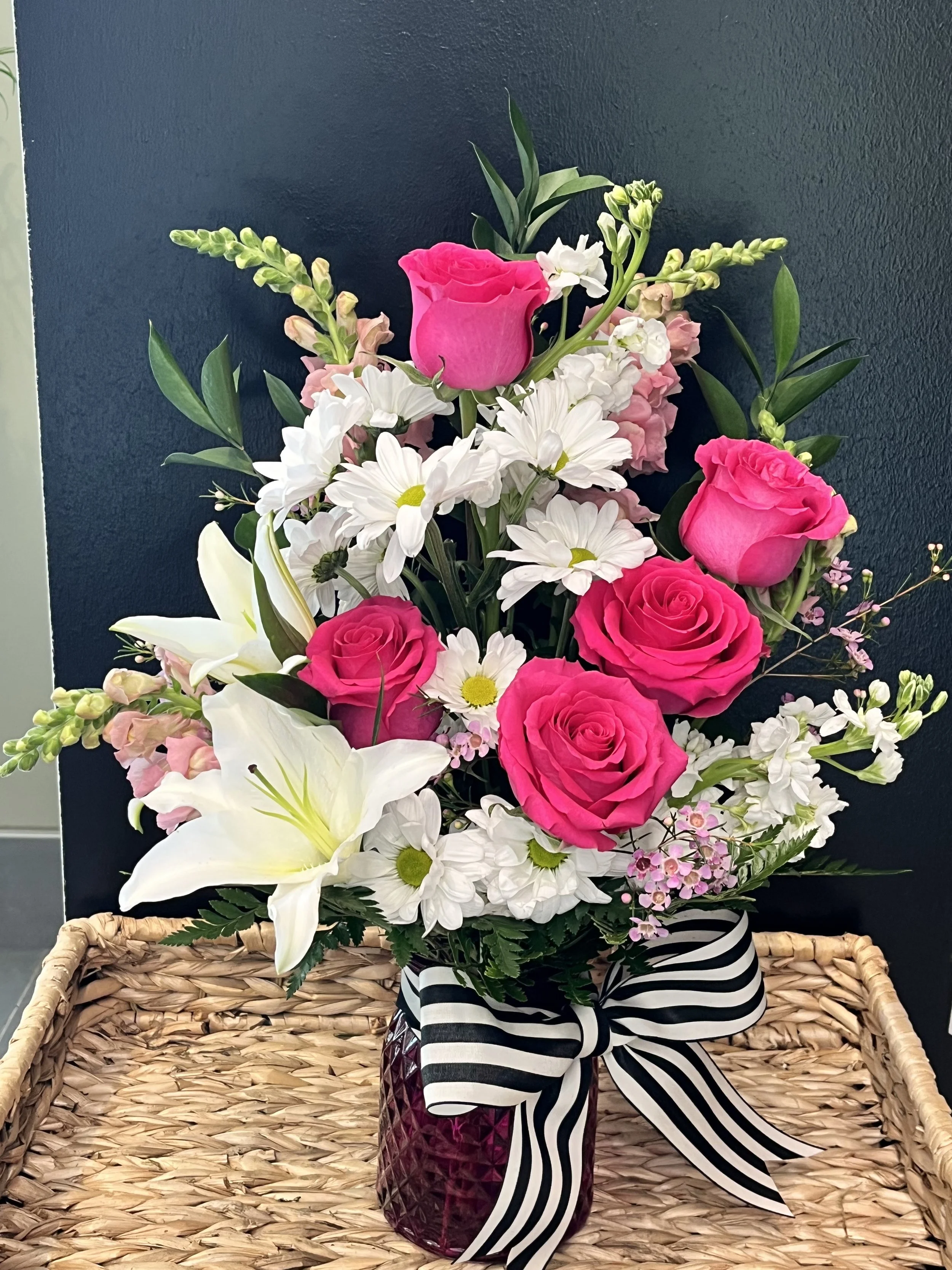 Bright pink roses, white lilies, daisies, and other mixed flowers in a glass vase with a black and white striped ribbon, placed on a wicker tray against a dark wall background.