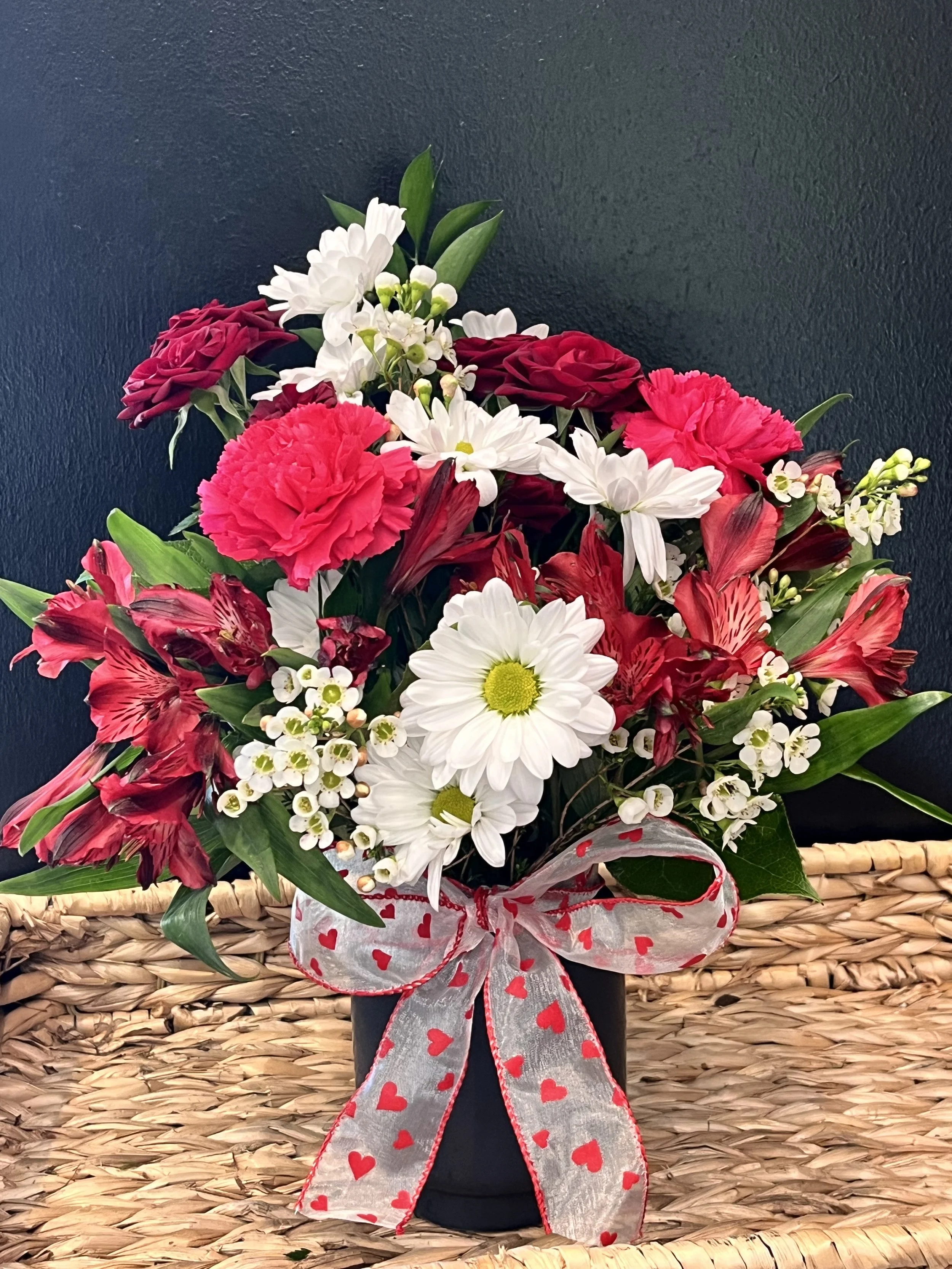 A bouquet of red, white, and pink flowers in a black pot, decorated with a ribbon with red hearts, placed in a woven basket against a dark background.