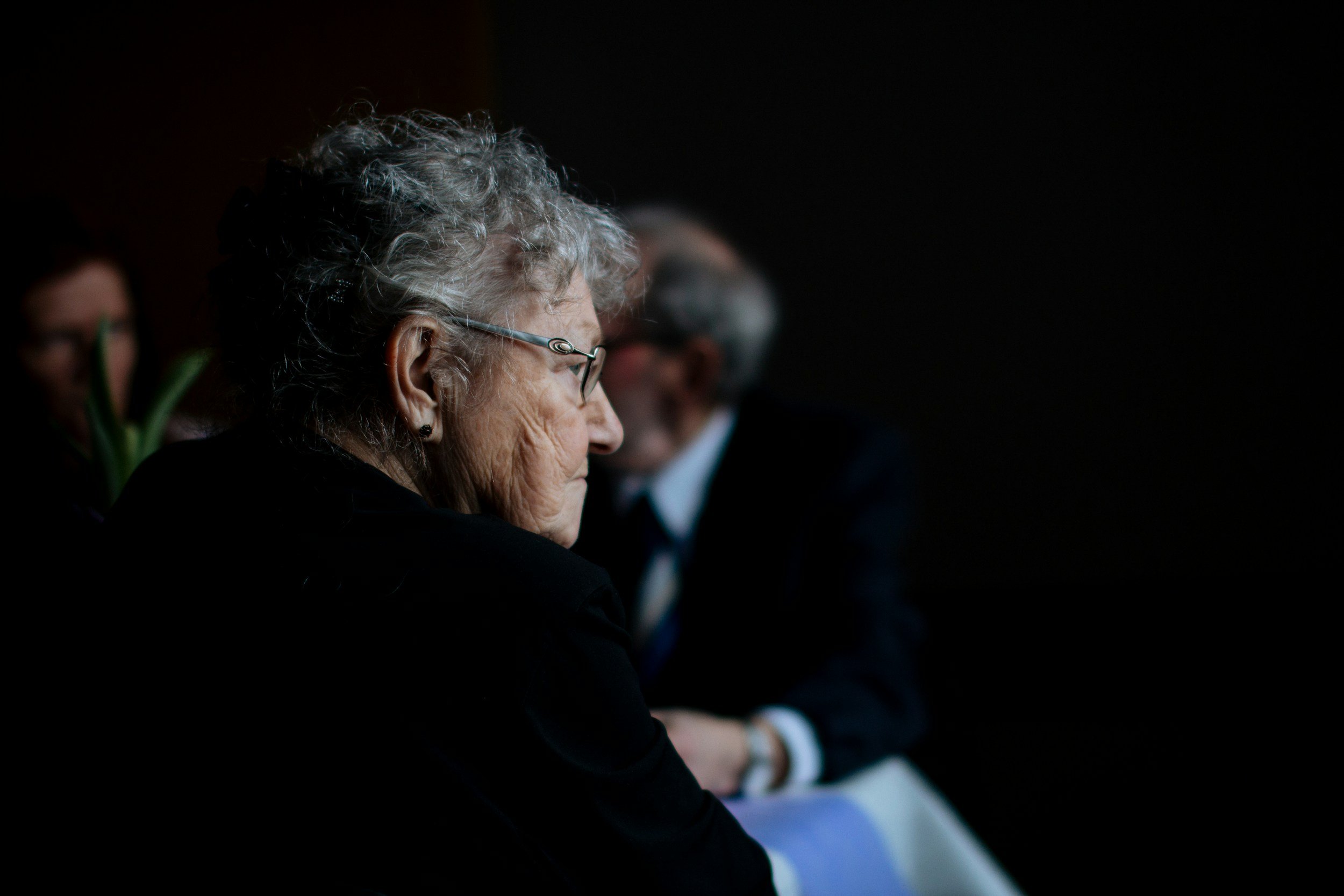 Side profile of an elderly woman with curly gray hair wearing glasses, sitting in darkness. Other people are blurred in the background.