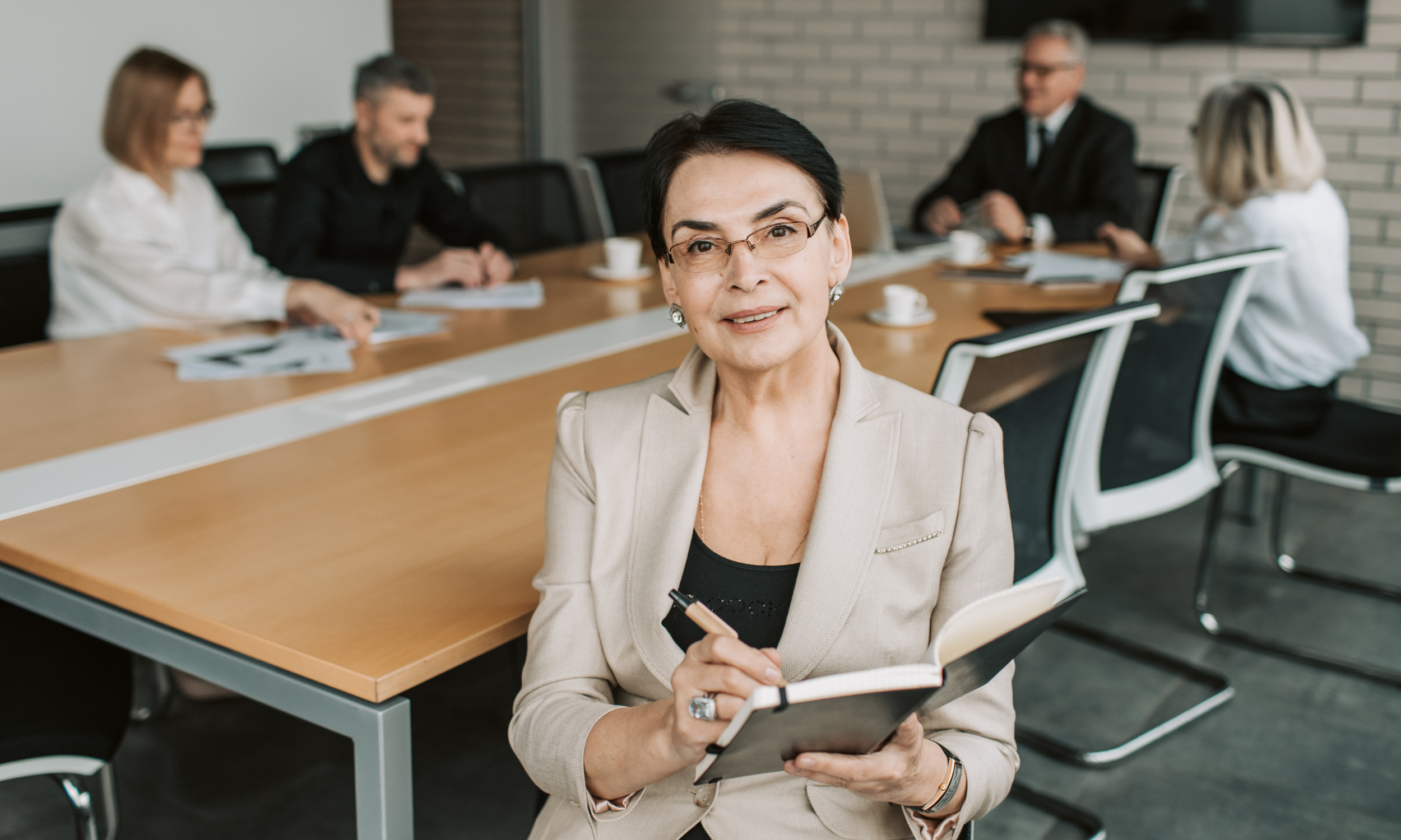 A confident woman with short black hair and glasses sitting at a conference table, holding a notepad and pen, with colleagues working in the background.