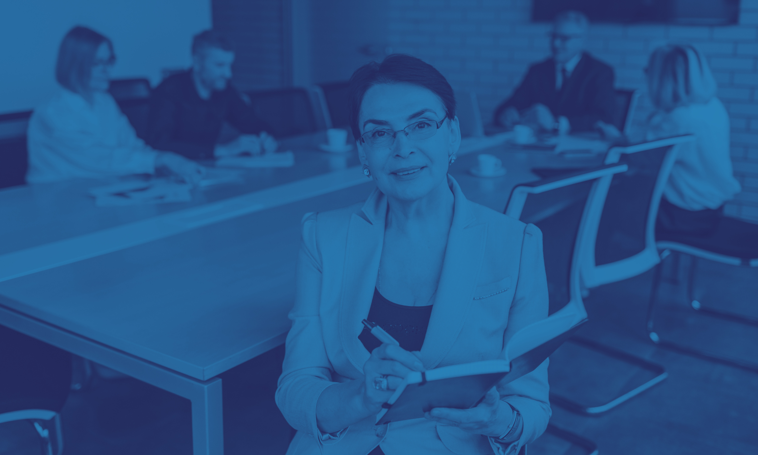 A woman wearing glasses and a blazer sitting at a conference table with a group of people in a meeting room.