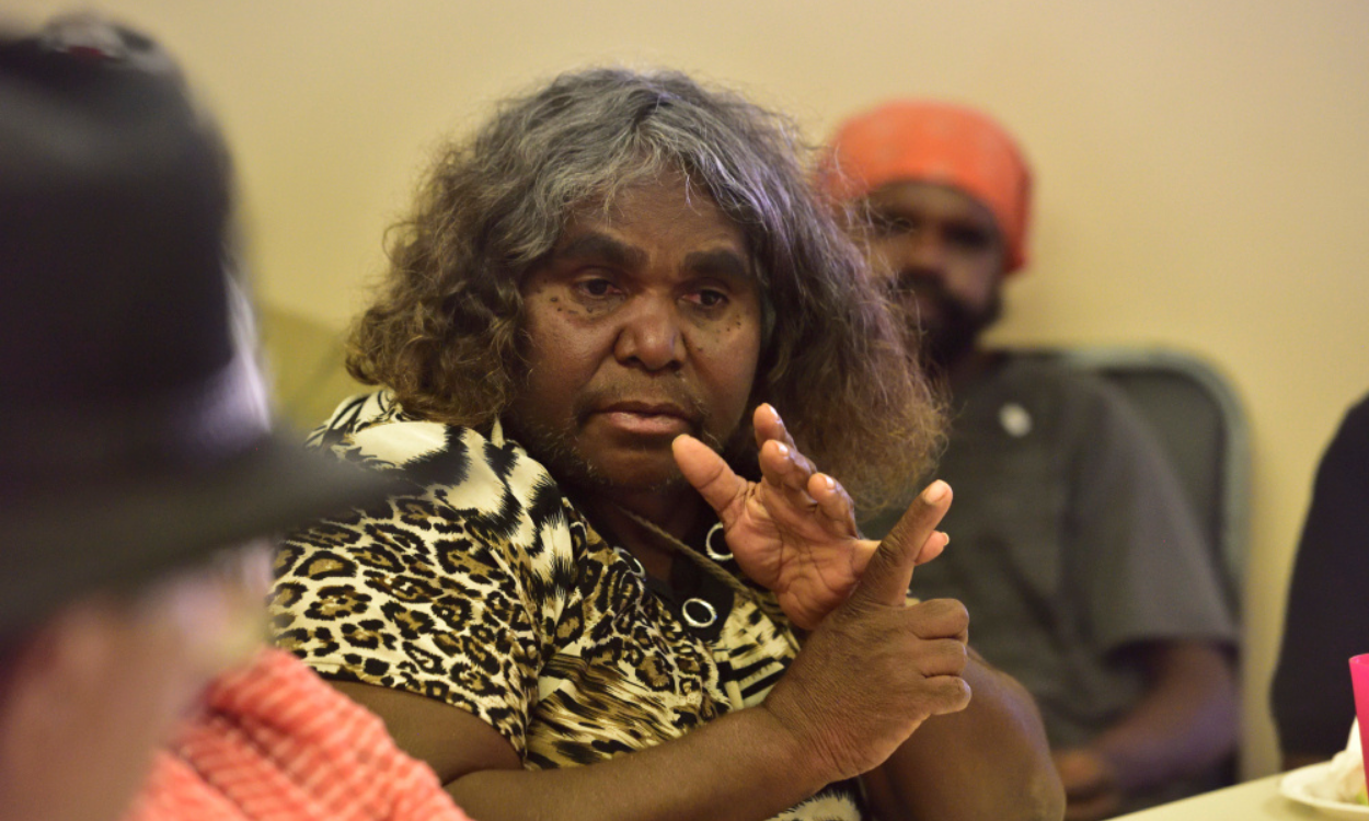 A person with long, curly hair and dark skin is speaking, gesturing with their right hand, in a meeting or discussion, wearing a leopard print shirt. Other people are visible in the background and foreground.
