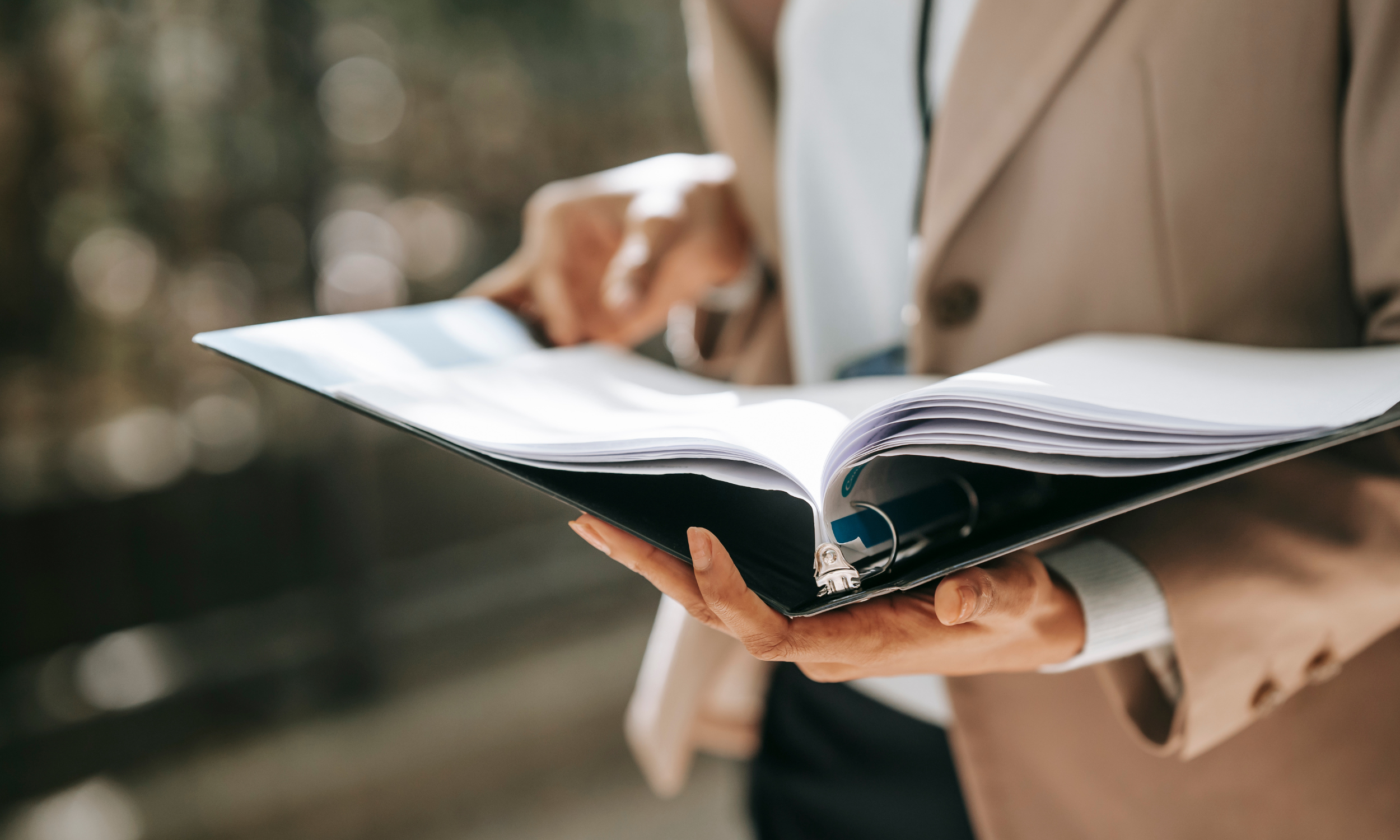 A person in a tan coat holds an open binder with papers, while another person looks at their phone in an outdoor setting with blurred background.