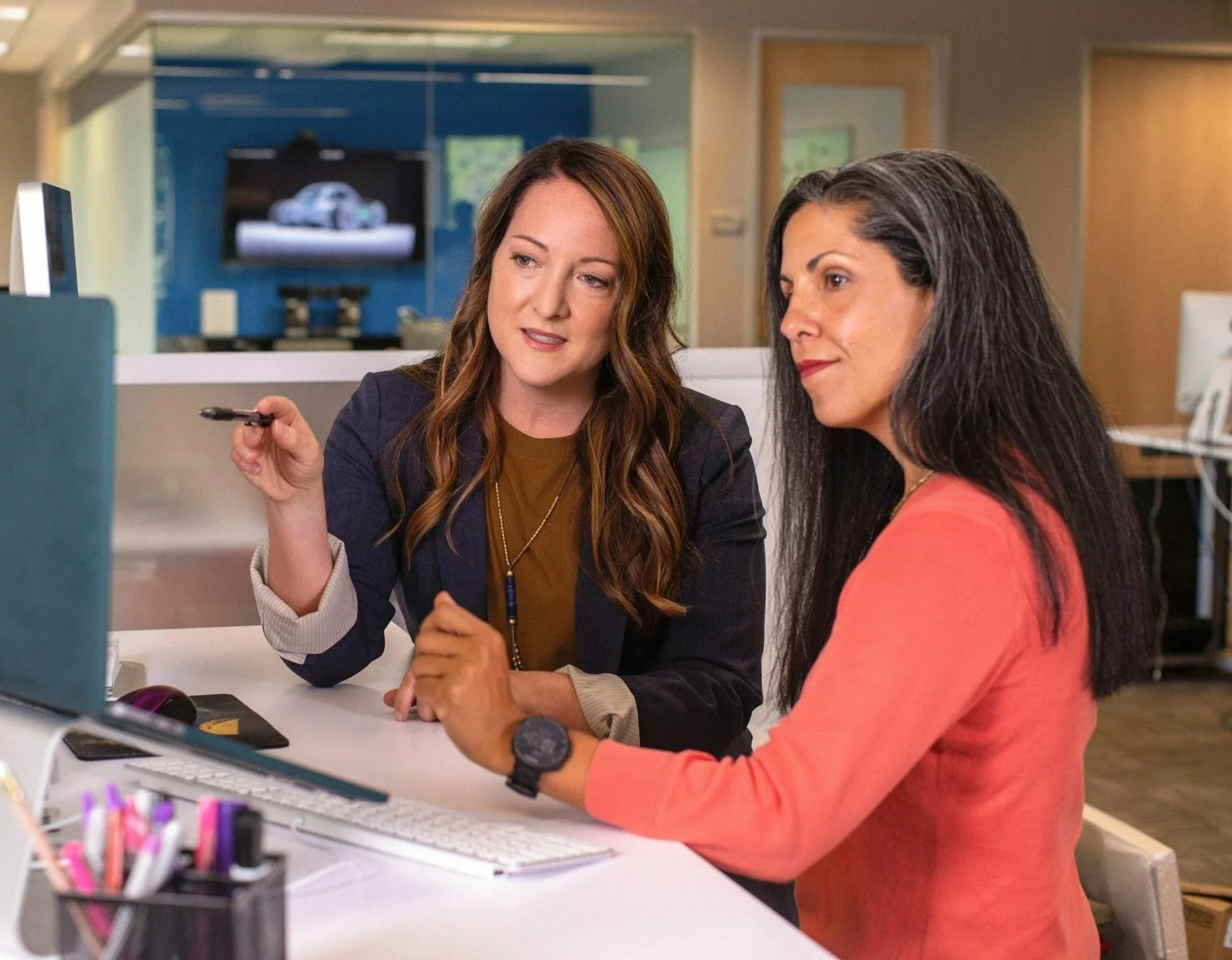 Two women sitting at a desk in an office, engaged in a conversation, with office supplies and a computer in the foreground.