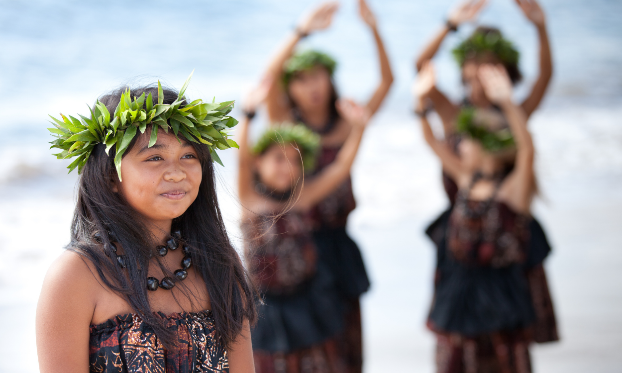 Young girl wearing a leaf crown and traditional Polynesian clothing standing on beach, with three women in the background performing a hula dance.