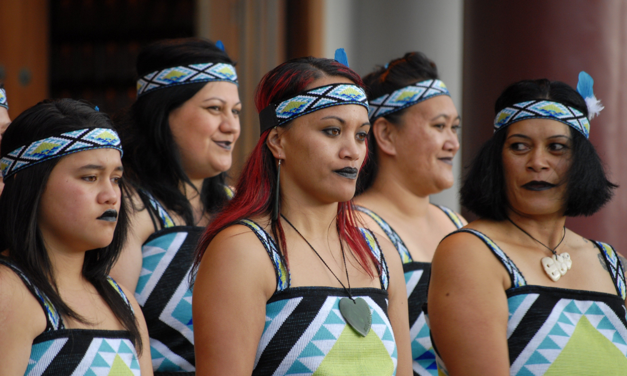 A group of Indigenous women wearing traditional patterned tops and headbands, participating in a cultural ceremony or gathering.