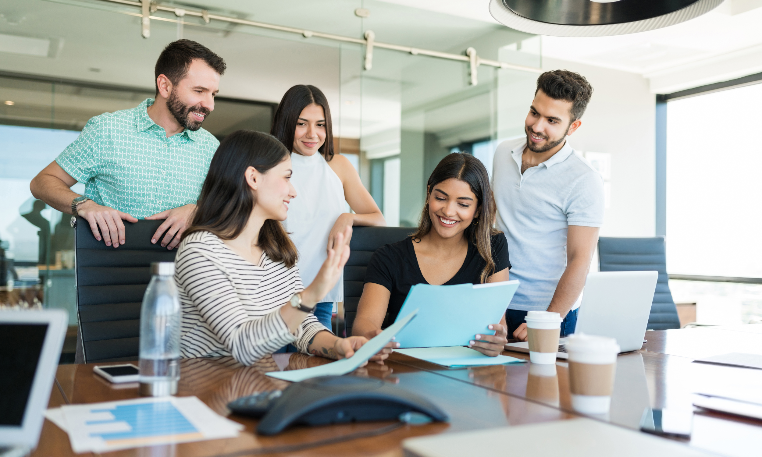 Group of five young professionals collaborating around a conference table in a modern office, discussing and reviewing documents with laptops and coffee cups.