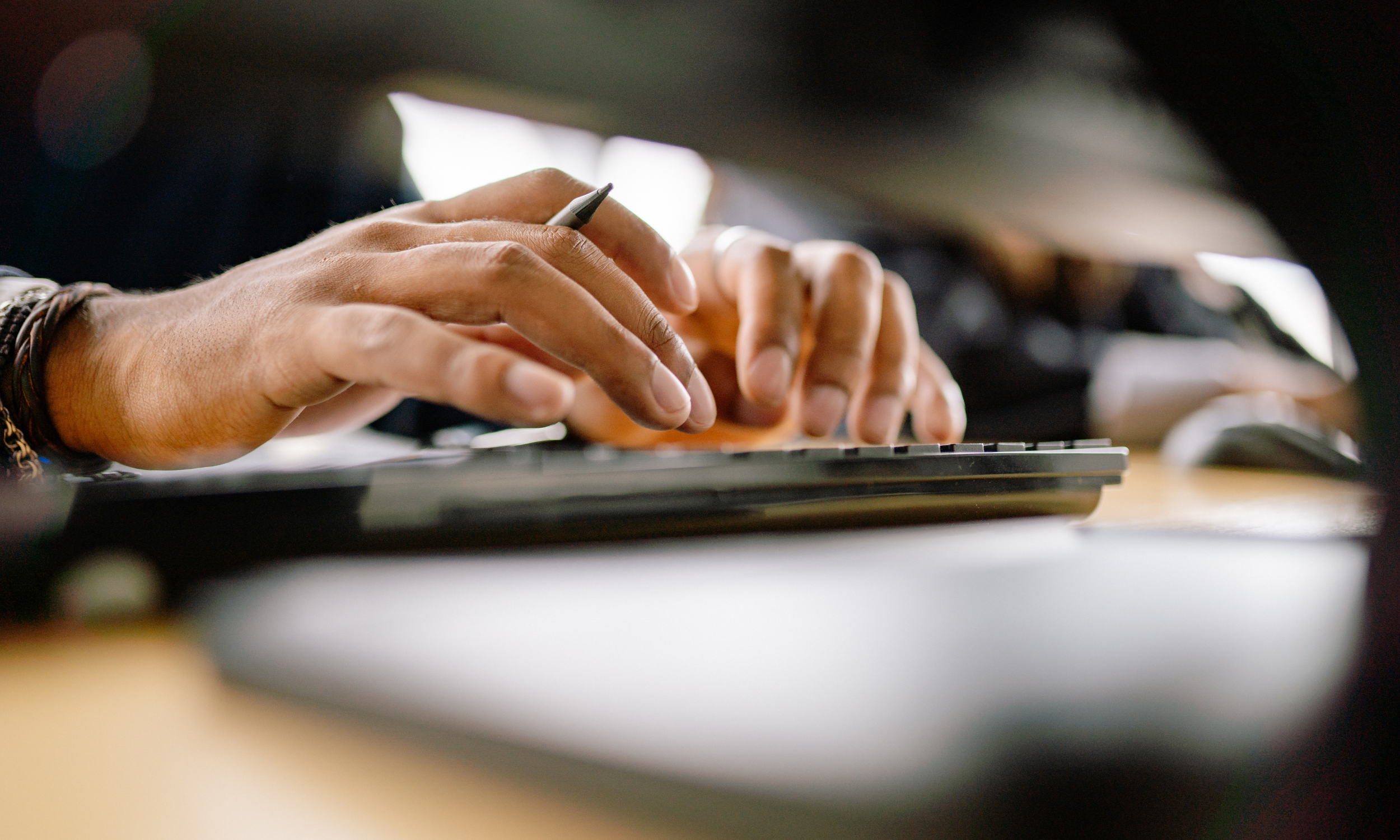 Close-up of hands typing on a keyboard, with focus on the fingers and wrist accessories, in a dimly lit environment.