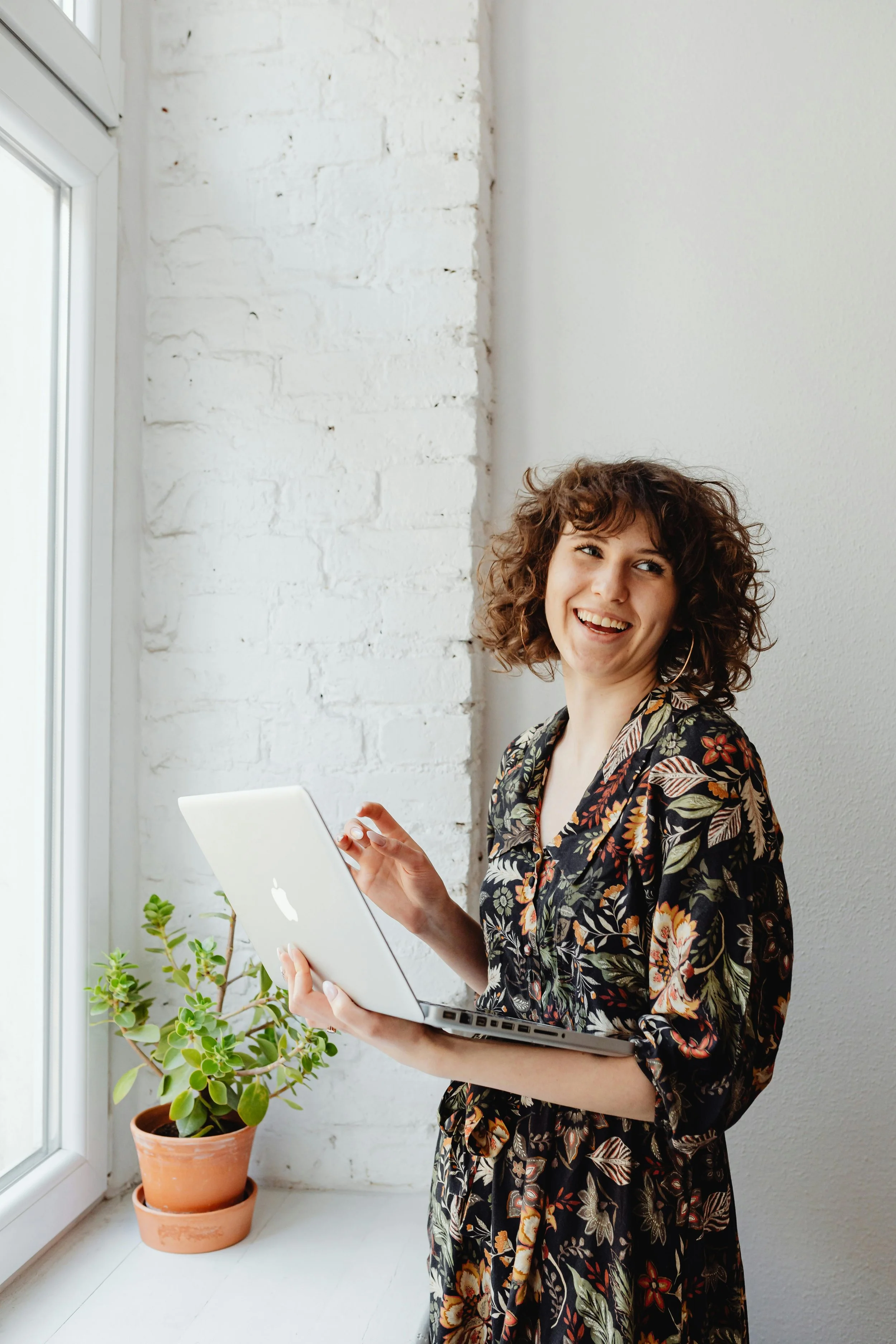 A smiling woman with curly hair holding a MacBook laptop near a window with white brick wall and plants nearby.