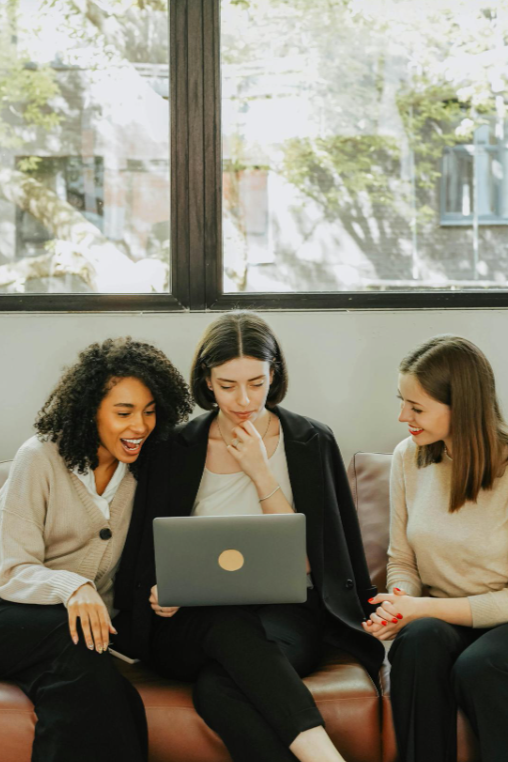 Three women sitting on a couch looking at a laptop and smiling.
