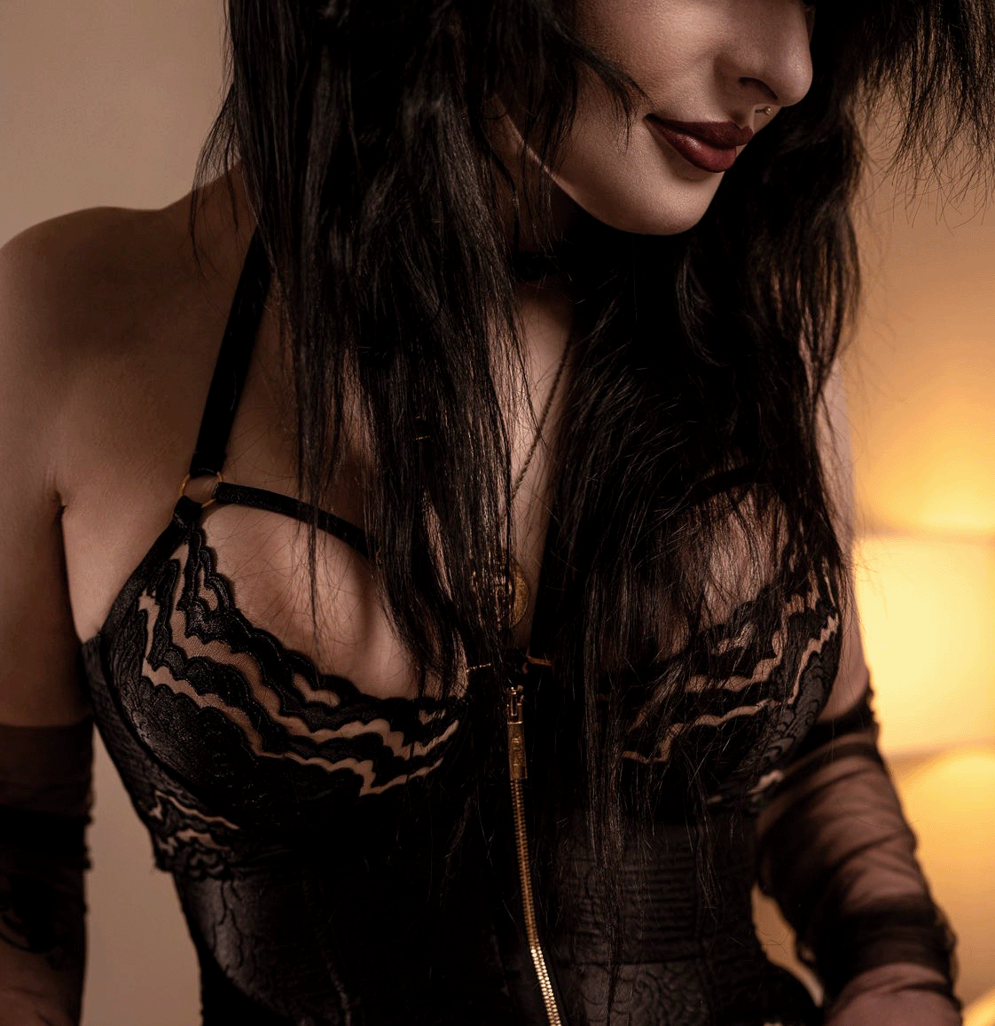 Close-up of a woman with long dark hair, wearing black lace lingerie and a gold necklace, smiling softly in a warm-lit room.