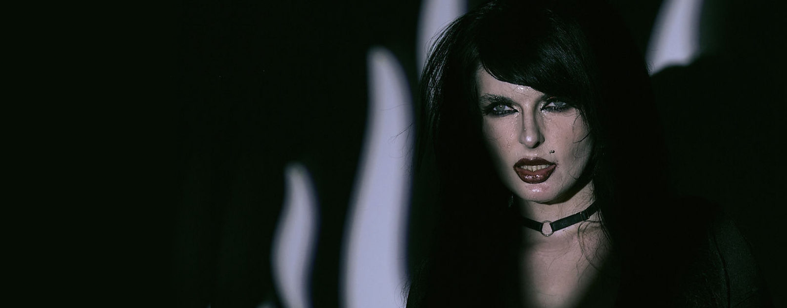 Close-up of a woman with dark hair, makeup, and a black choker, in a dark setting with a serious expression.