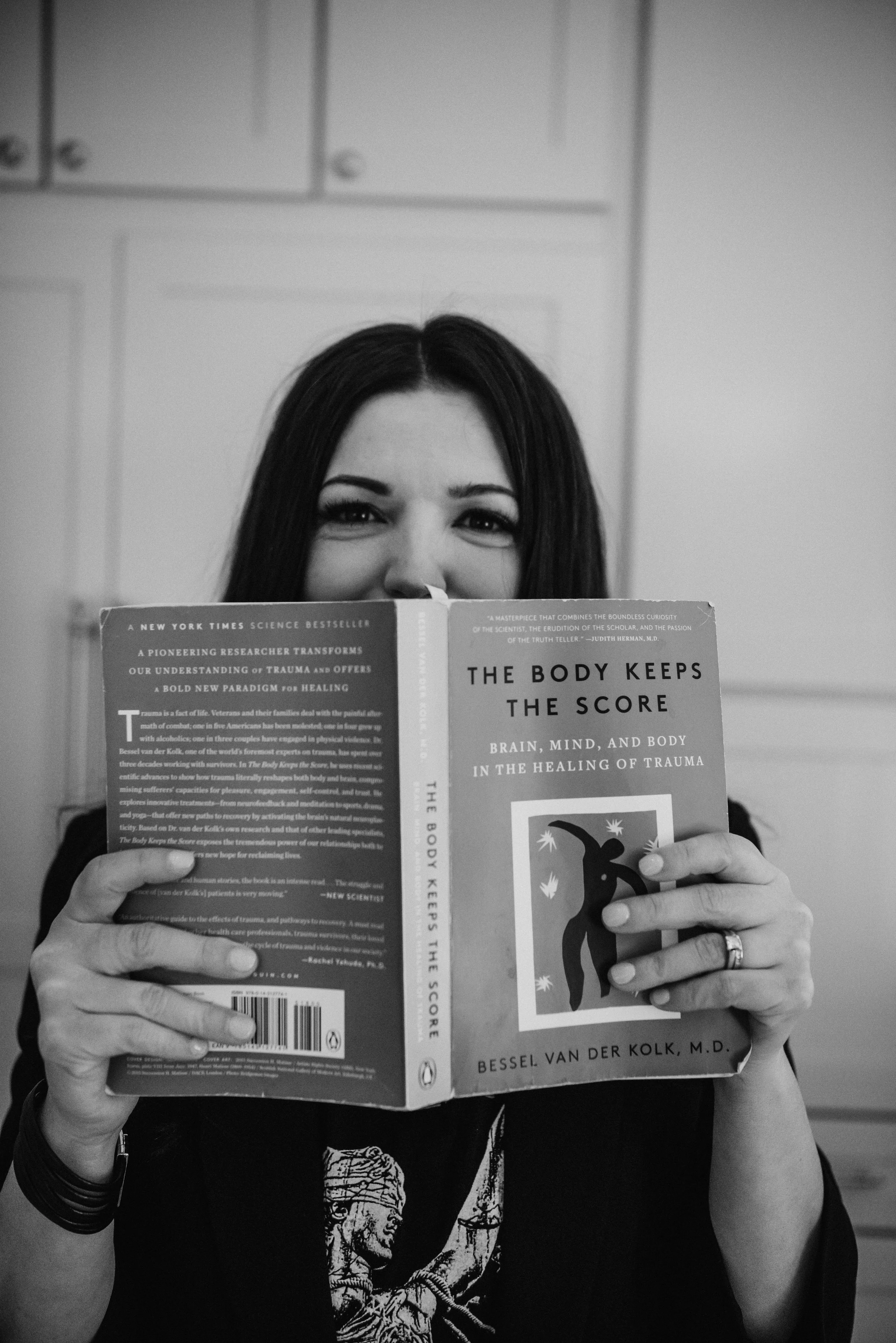 A woman with long dark hair holding a book titled 'The Body Keeps The Score' in front of her face, partially obscuring her face. She is smiling with visible eyes and eyebrows. The background shows a plain wall with paneling.
